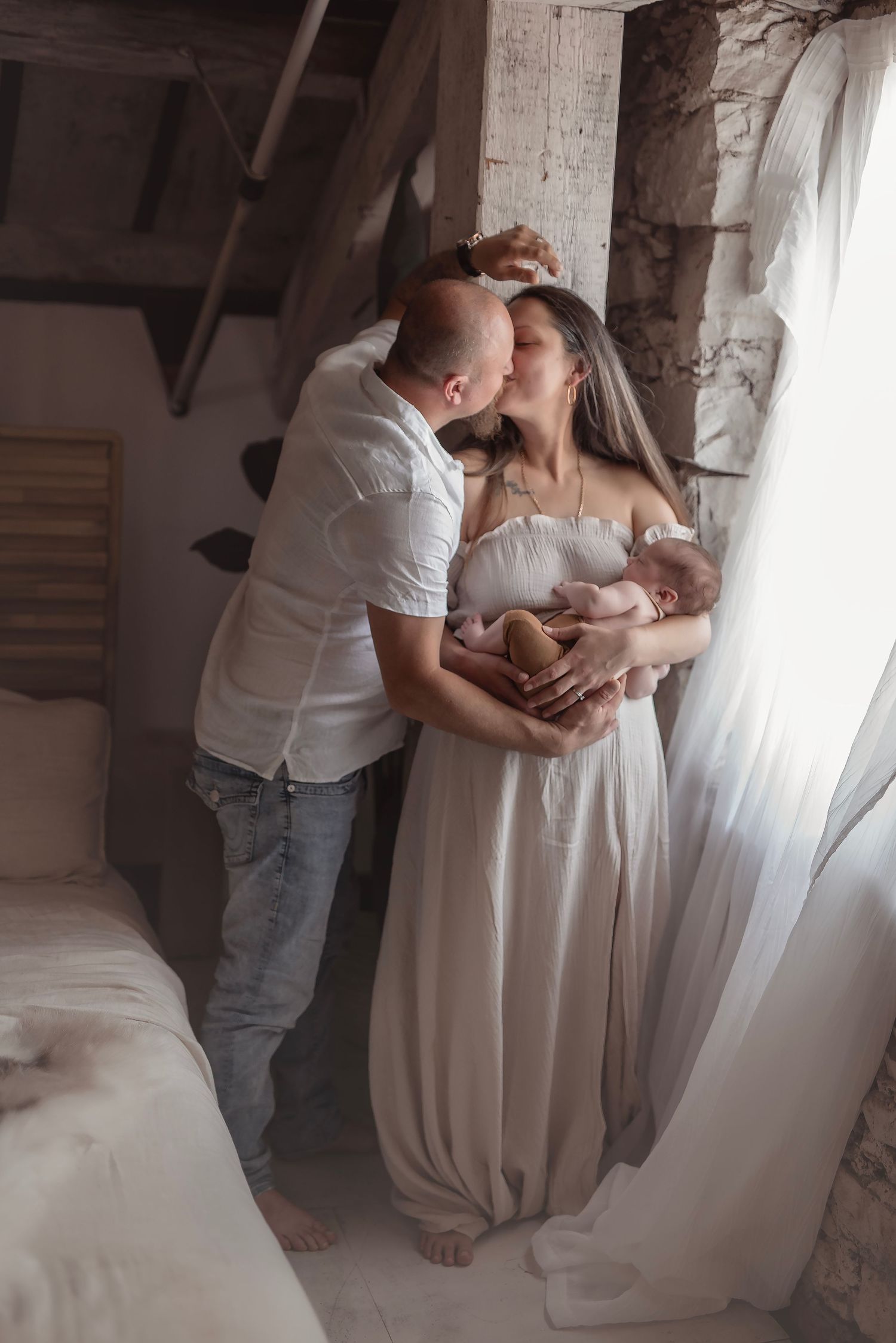 Couple kissing while mother holds newborn baby near window in soft-lit rustic bedroom setting.