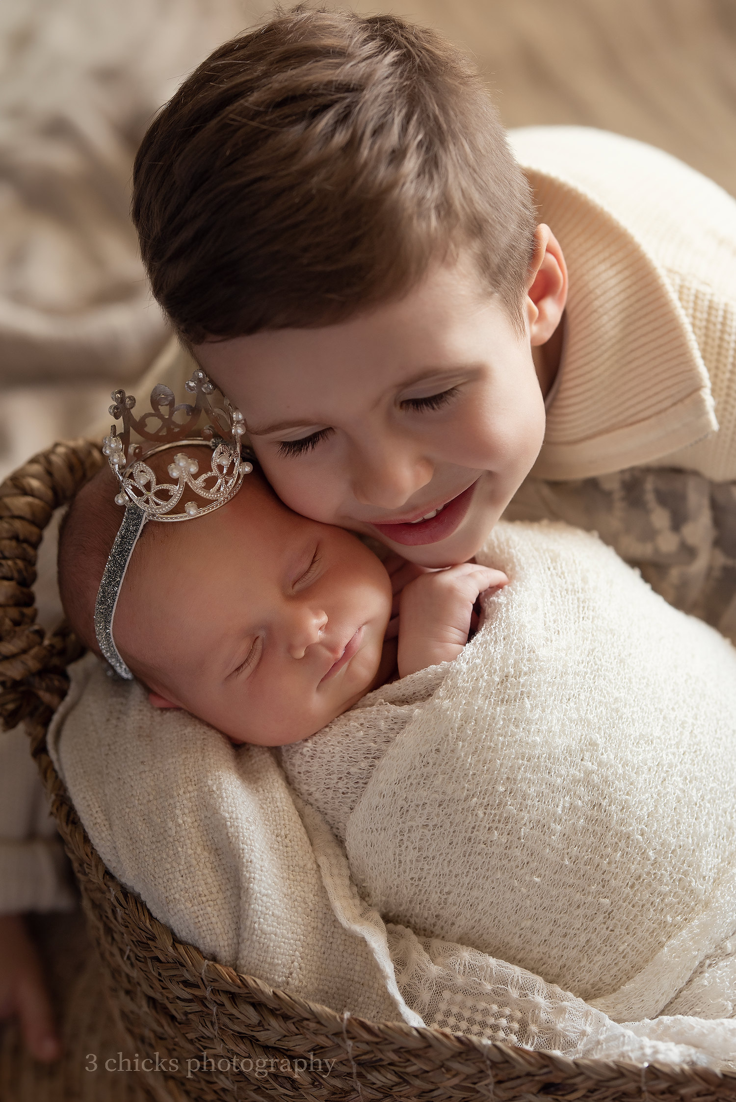 Older boy smiling while gently resting his chin on sleeping newborn baby wearing a small crown headband in a basket.