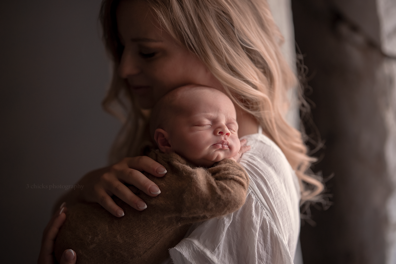 A mother tenderly holds her sleeping newborn baby wrapped in a brown swaddle against her shoulder.