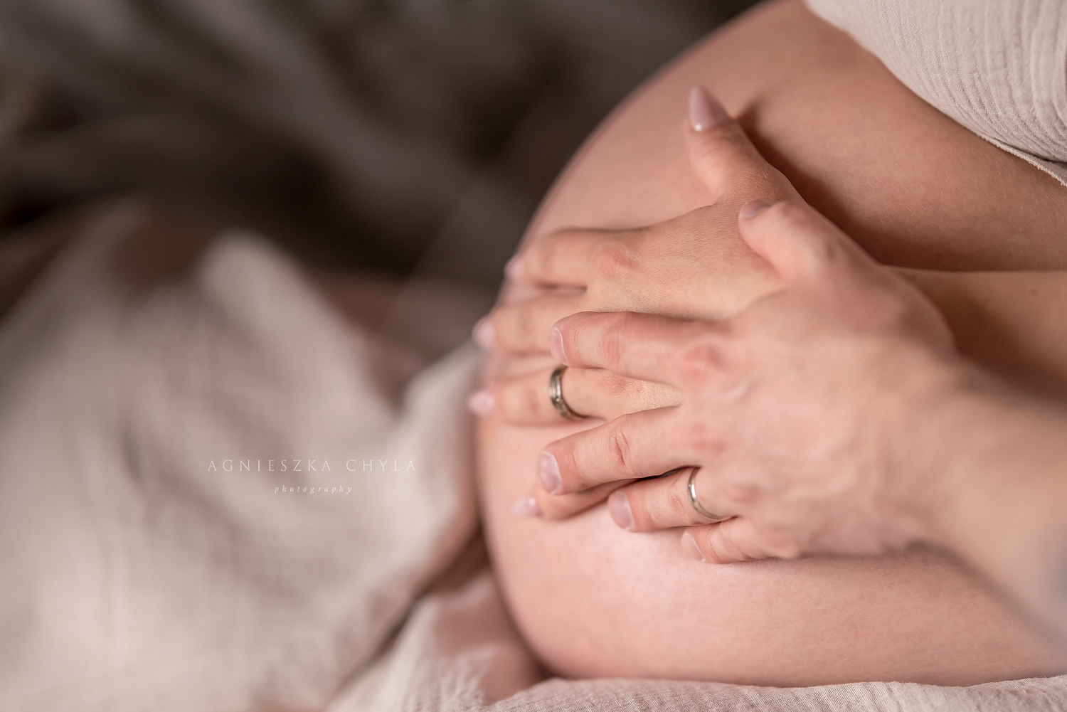 Close-up of a pregnant belly with two pairs of hands gently cradling the bump, one wearing a wedding ring.