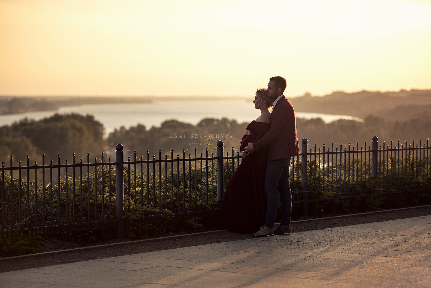 Couple embracing at sunset overlooking a scenic waterfront, silhouetted against a golden sky at a railed overlook.