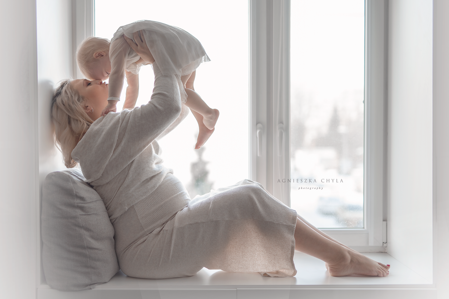 A mother sits by a bright window, lovingly lifting her baby in the air in a serene white room.