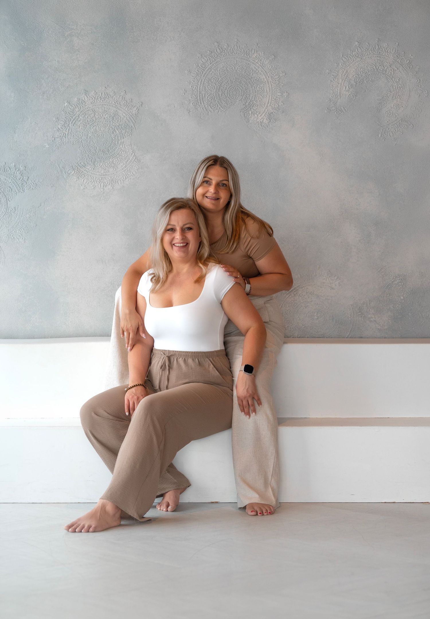 Two blonde women pose together in neutral tones, one seated and one standing, against a textured gray wall.