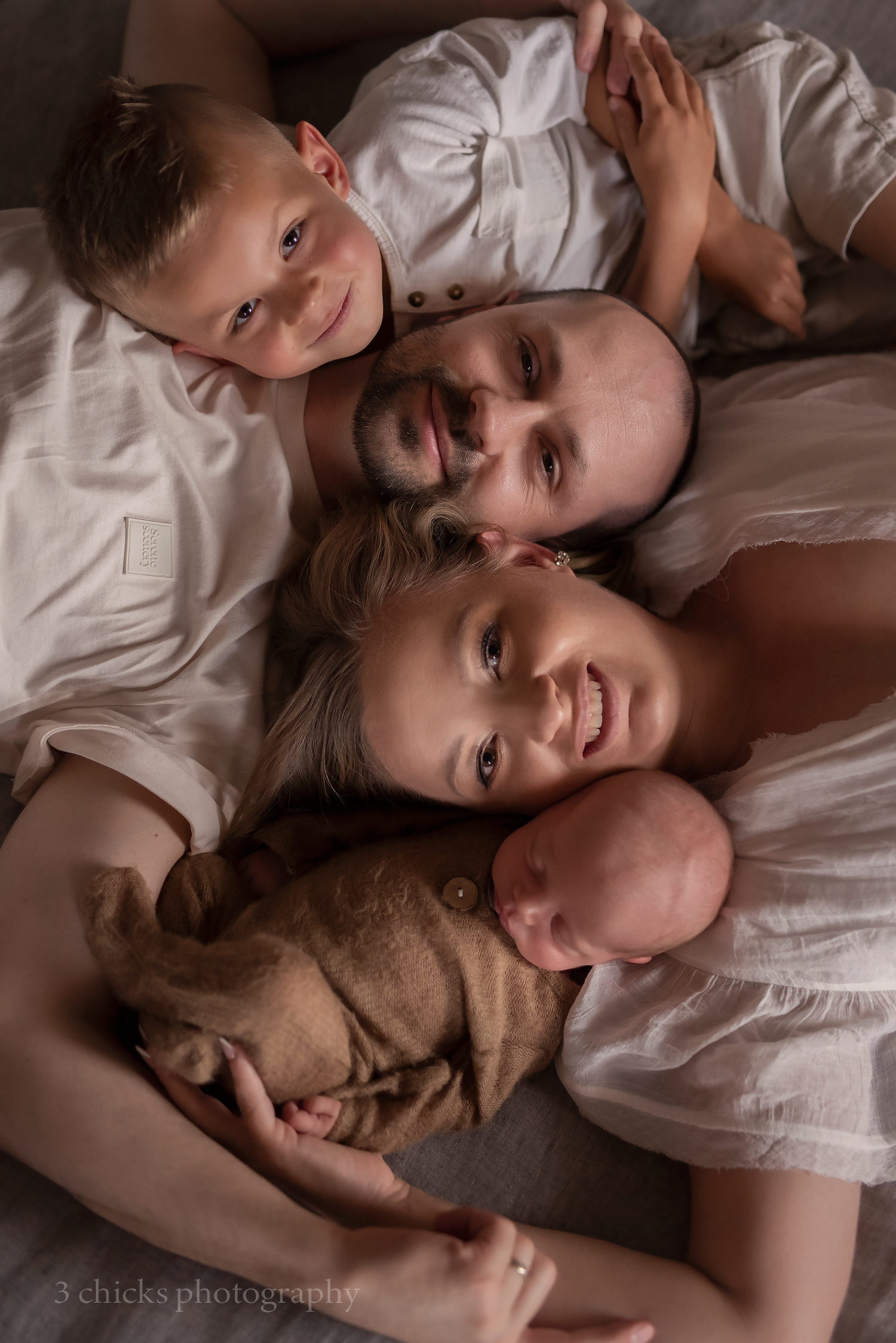 Family of four lying together, parents with toddler son and newborn baby, wearing white, captured by 3 Chicks Photography.