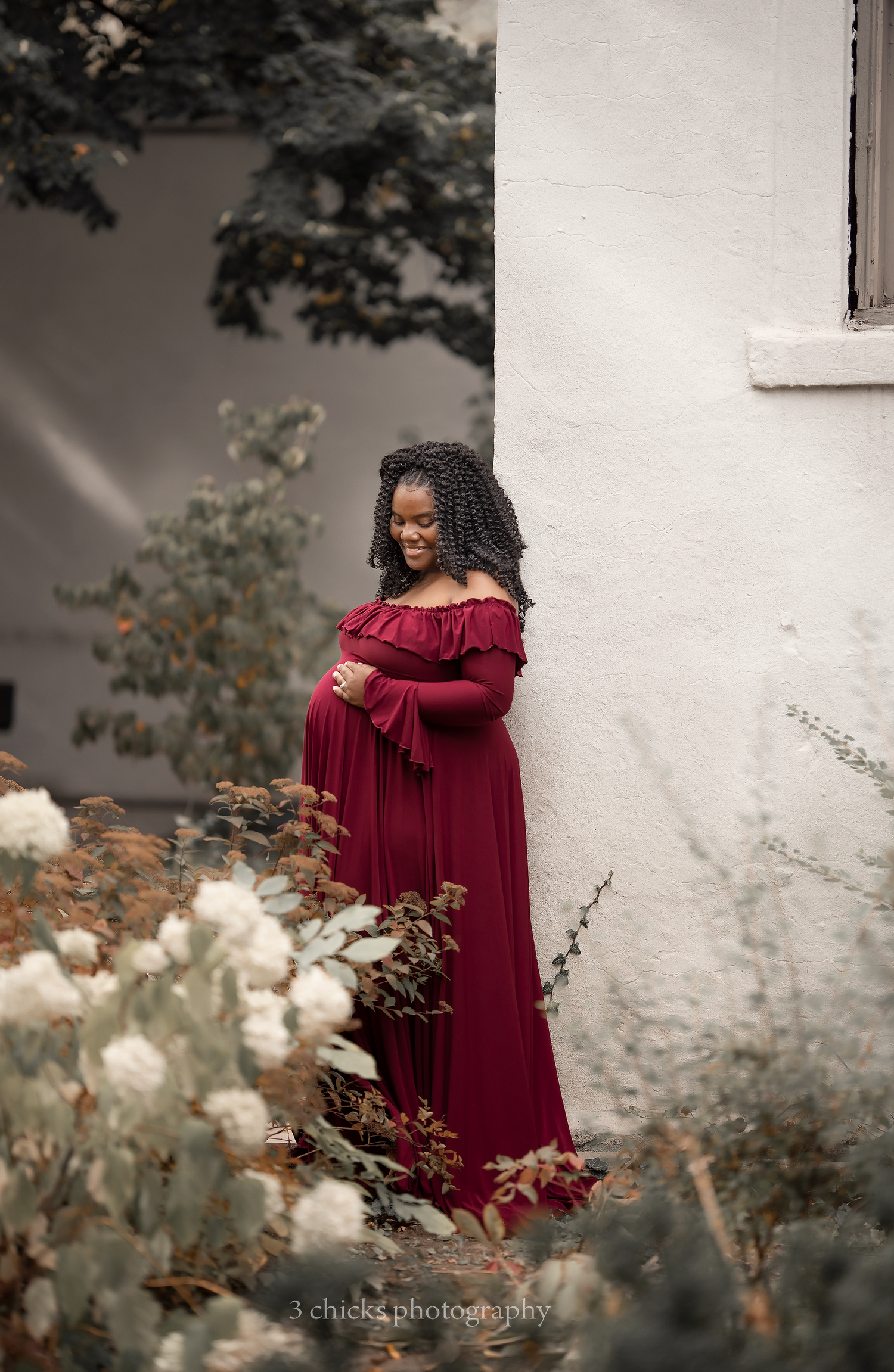 Pregnant woman in burgundy off-shoulder gown poses outdoors among white flowers by a white wall.