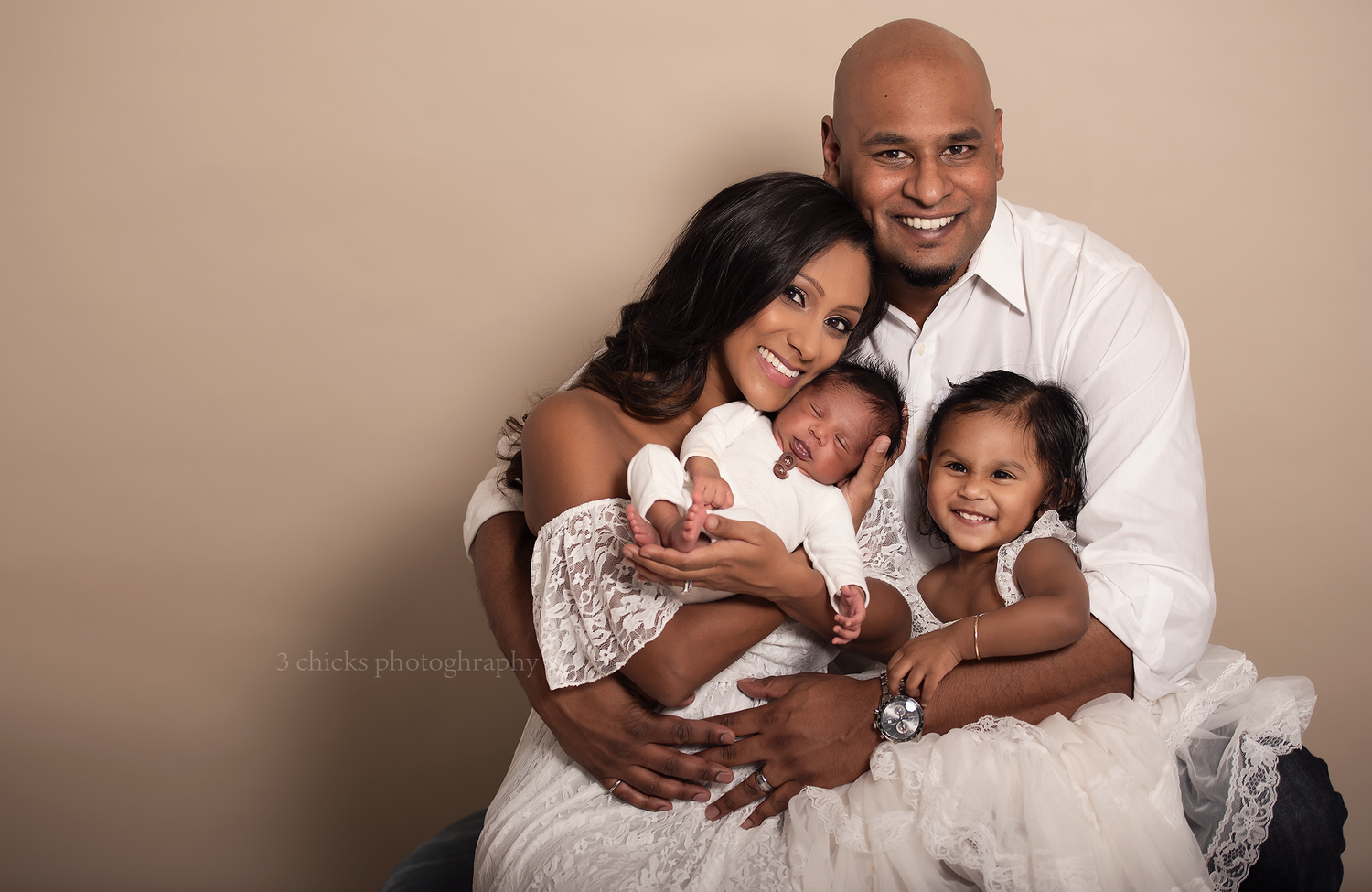 Happy family of four poses for a studio portrait, parents holding newborn baby and toddler daughter, dressed in white.