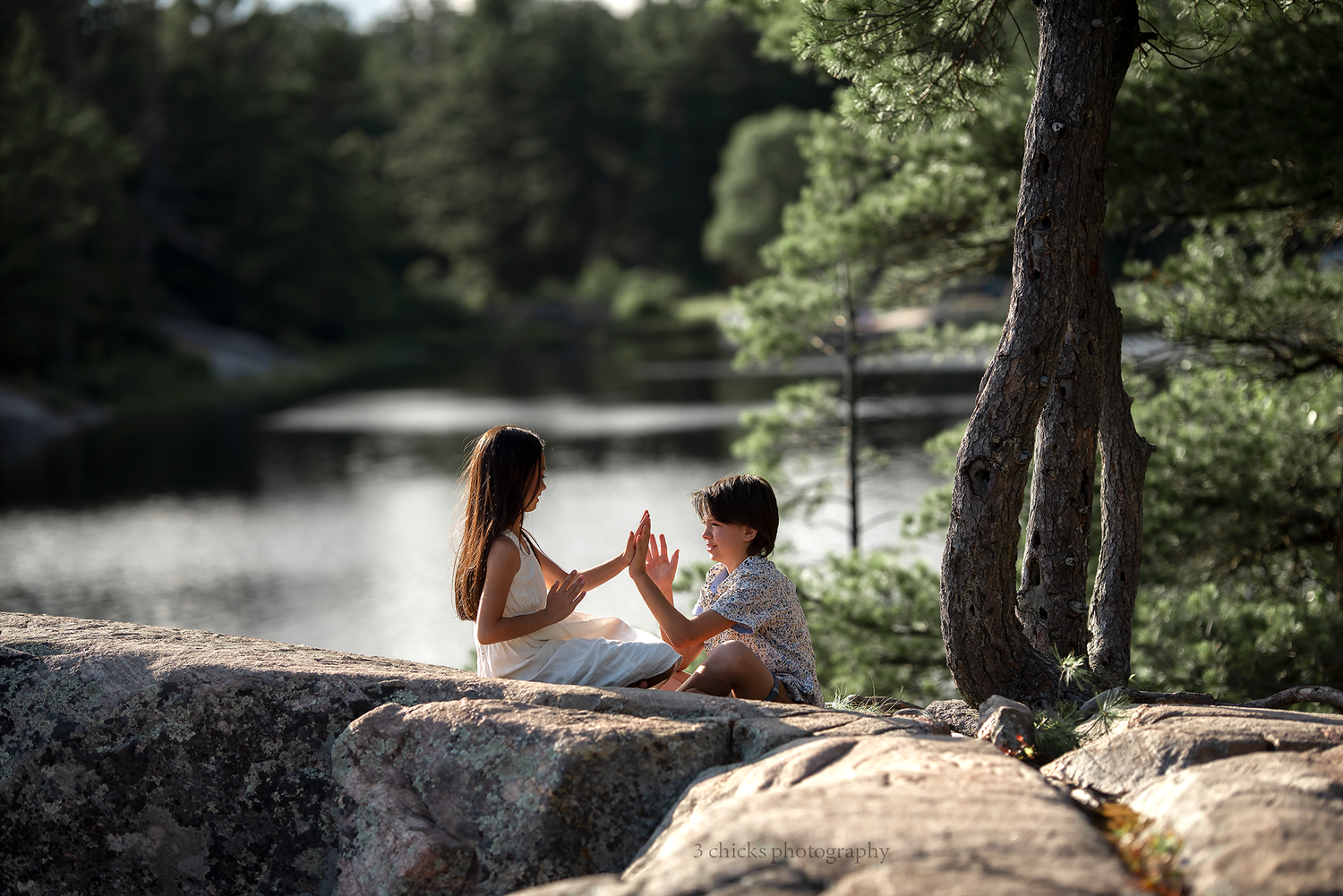Mother and child playing clapping games on rocky lakeside surrounded by pine forest.