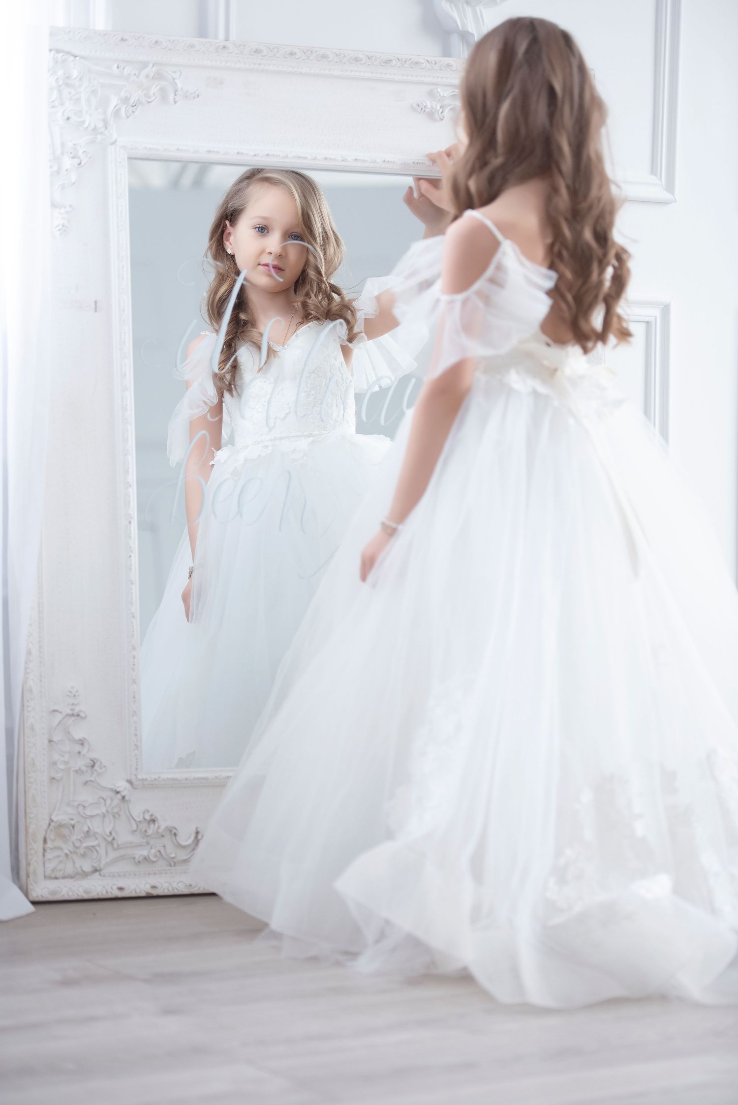Little girl in elegant white ball gown admiring herself in ornate mirror.