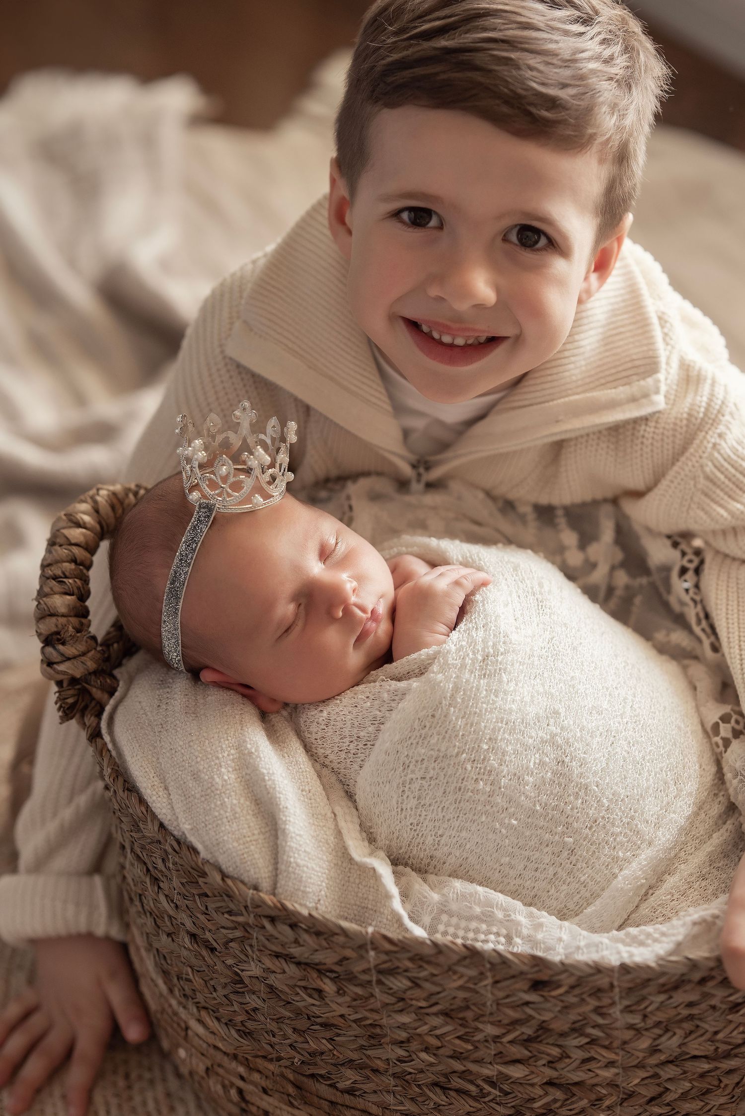 Smiling boy poses with sleeping newborn baby wearing a silver crown in a wicker basket wrapped in cream knit fabric.