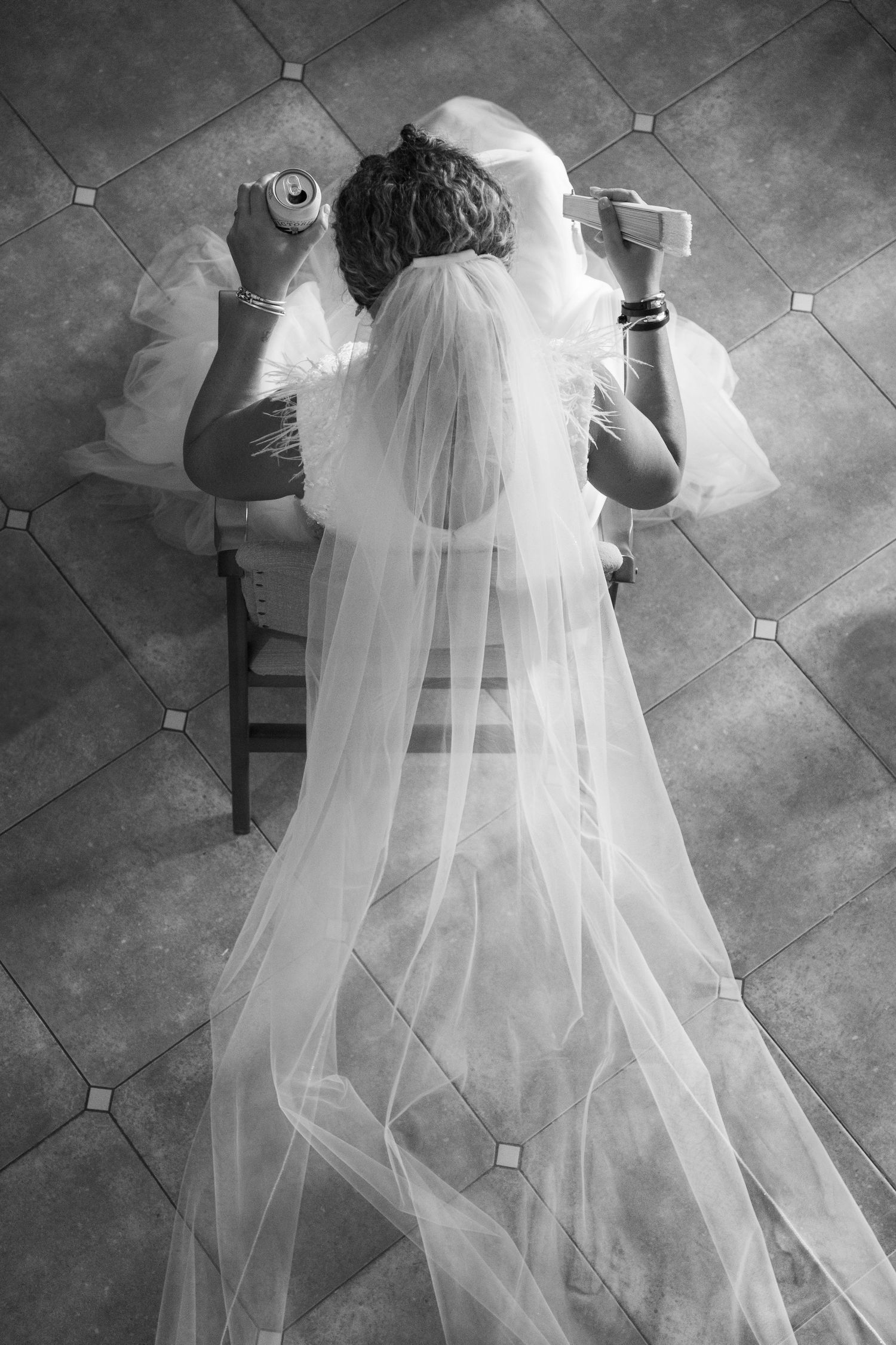 Fotografía artística cenital en blanco y negro de una novia sentada con su largo velo extendido, sosteniendo una cerveza y un abanico.