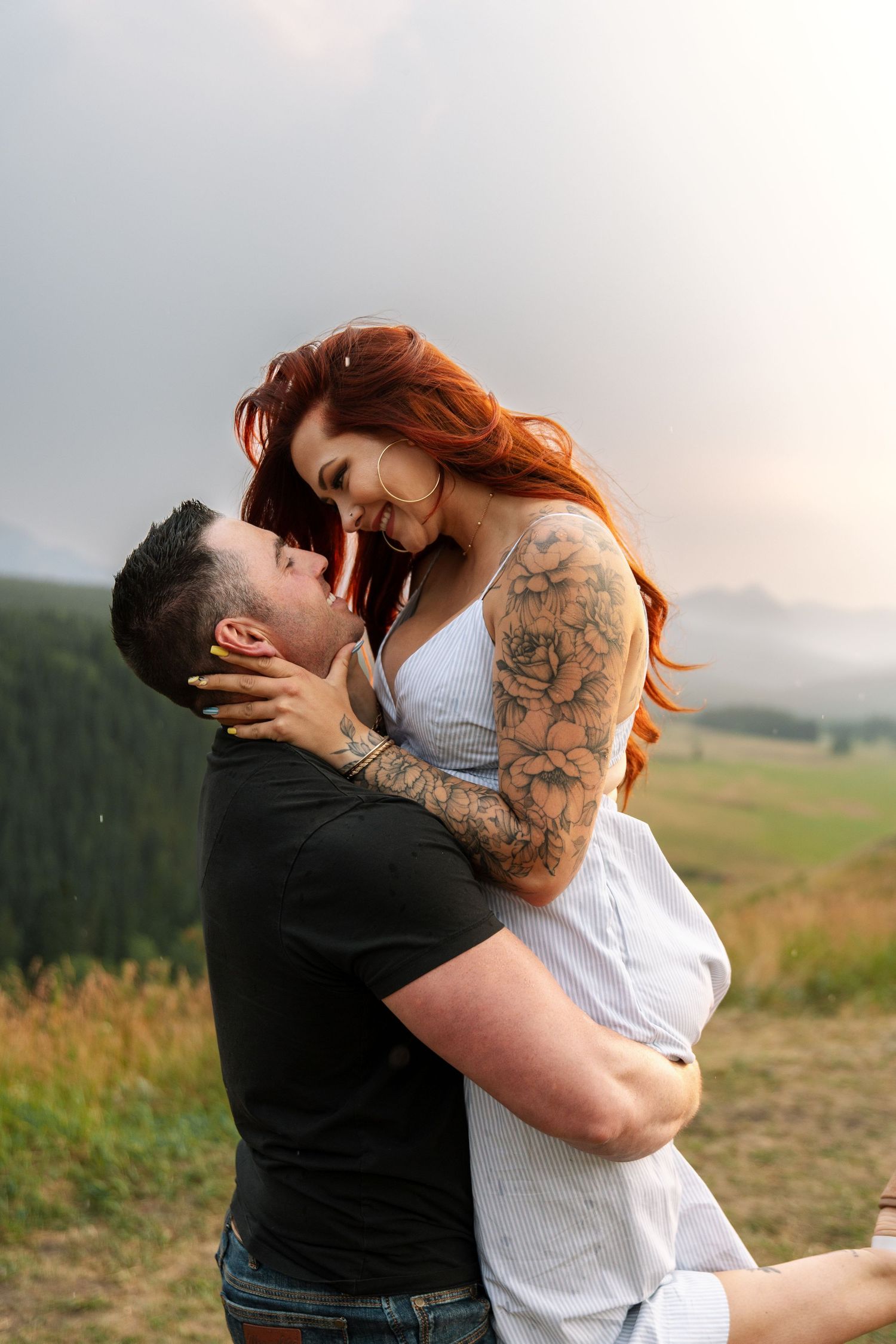 Man lifting woman with red hair and tattoos in a romantic outdoor mountain setting under a cloudy sky.