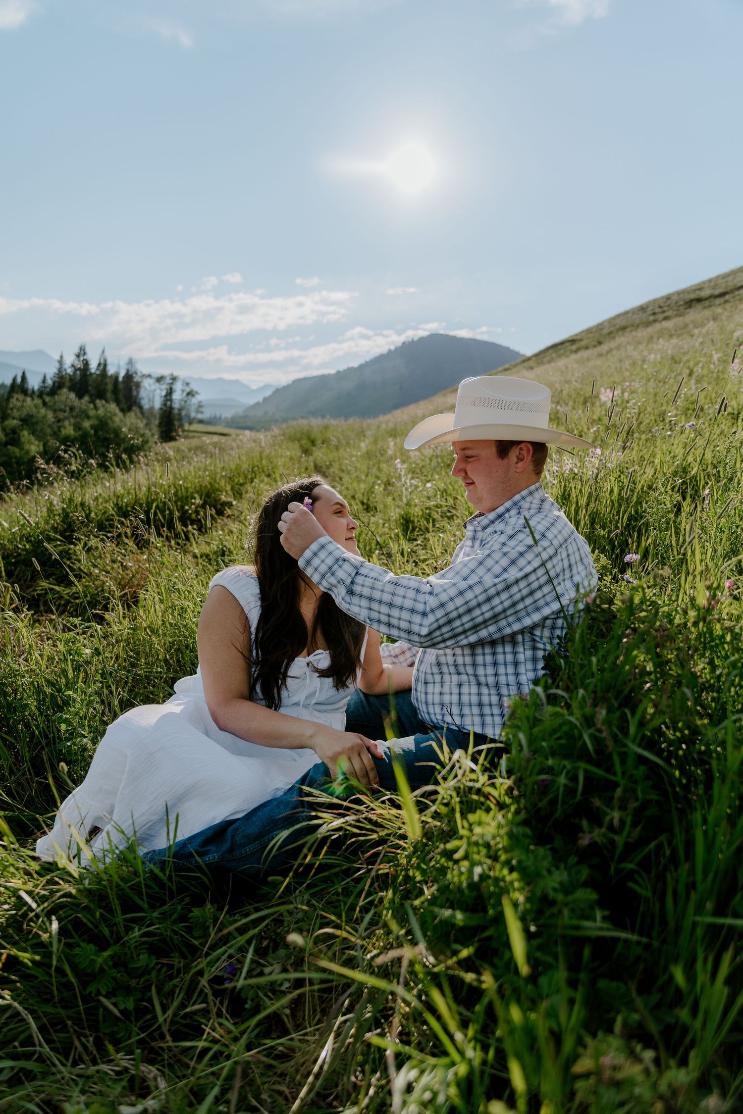 Couple sitting in lush green mountain meadow, man in cowboy hat tenderly touches woman's hair on sunny day.