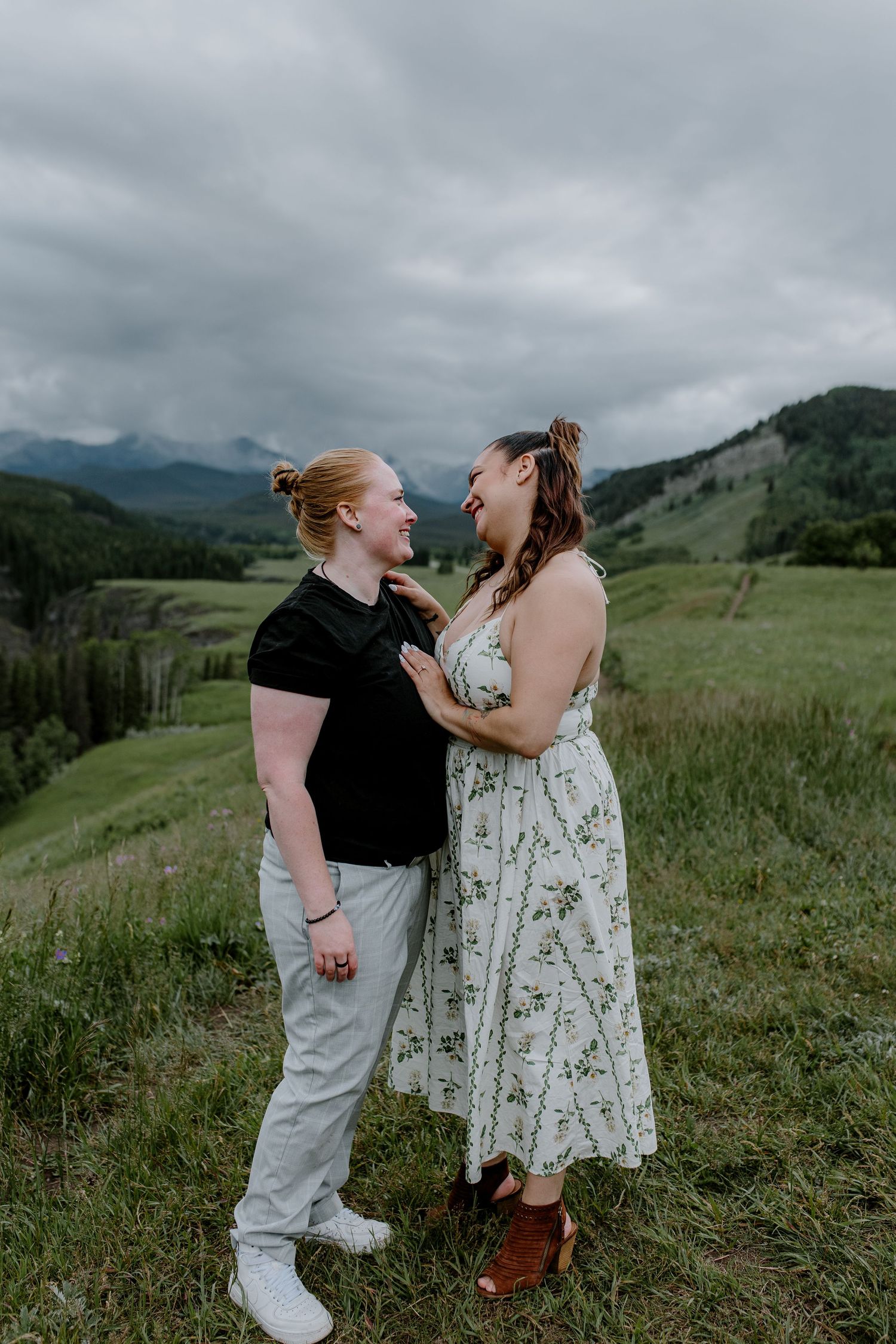 Smiling couple gazing at each other on a green hillside with dramatic cloudy mountain backdrop.