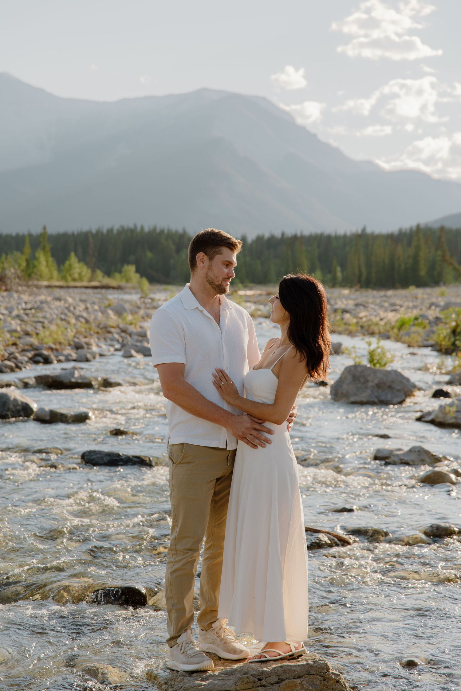 Couple embracing on rocky river with mountain forest backdrop at golden hour.