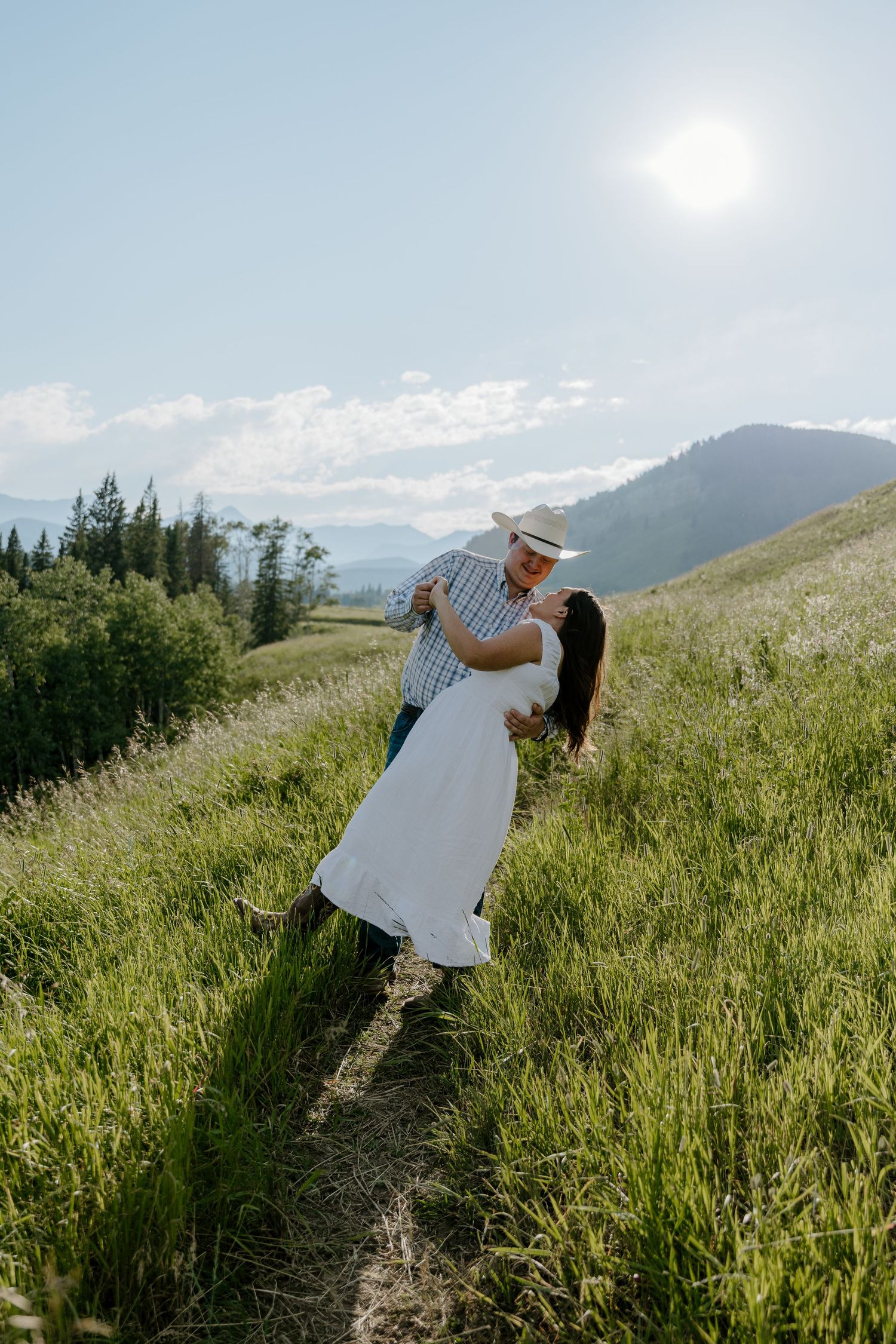 Couple dancing in a sunny green meadow with mountain views, man in cowboy hat dips woman in white dress.