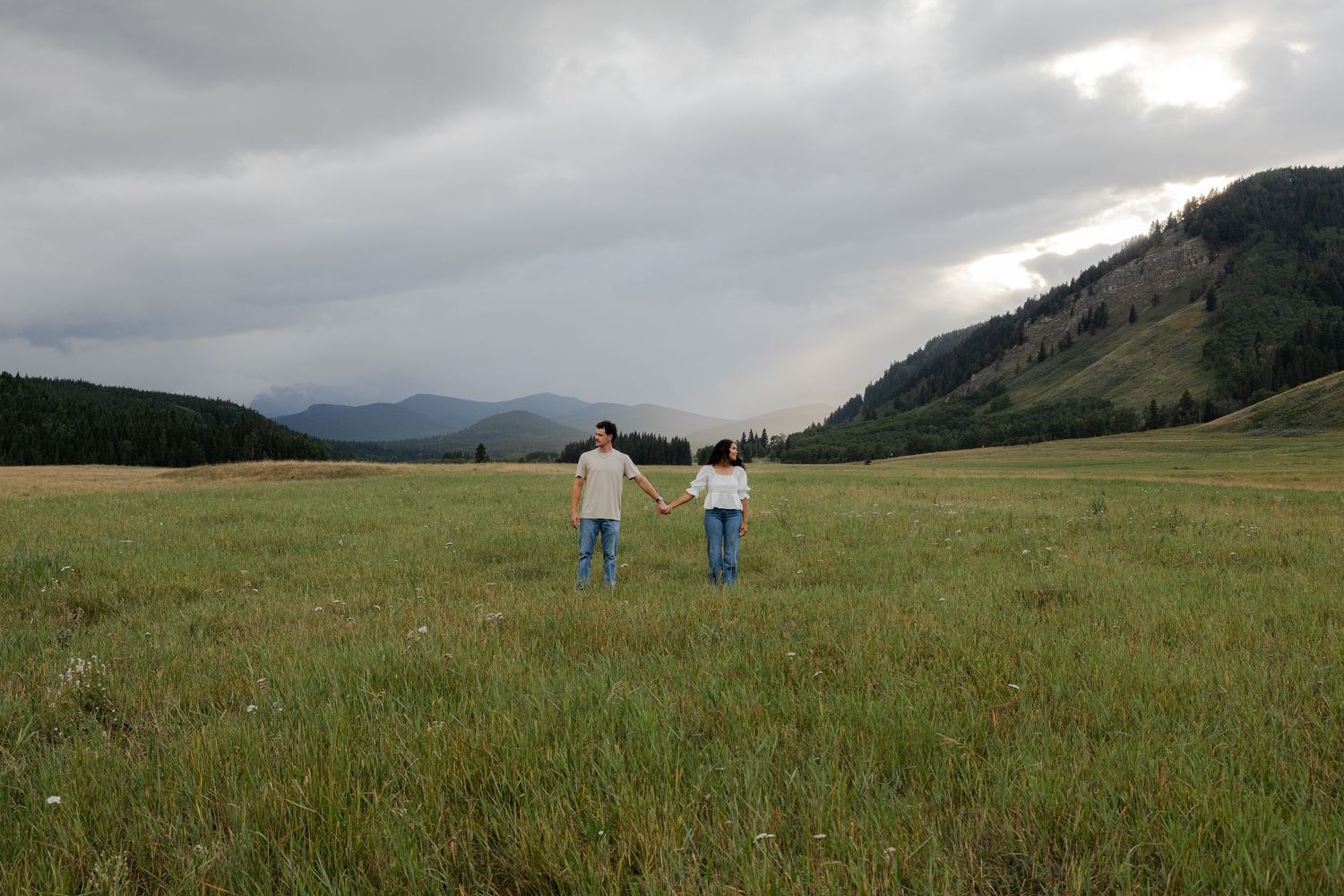 Couple holding hands in a vast green meadow with dramatic stormy skies and mountain backdrop.