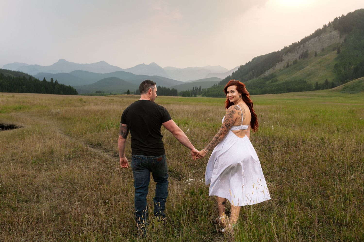 Couple holding hands walking through a lush green mountain meadow, woman in white dress with red hair, misty peaks behind.