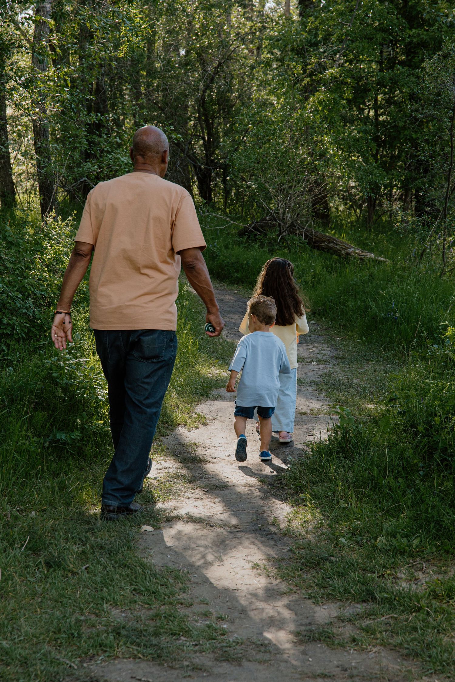 An adult and two children walk along a forest path surrounded by lush green trees on a sunny day.