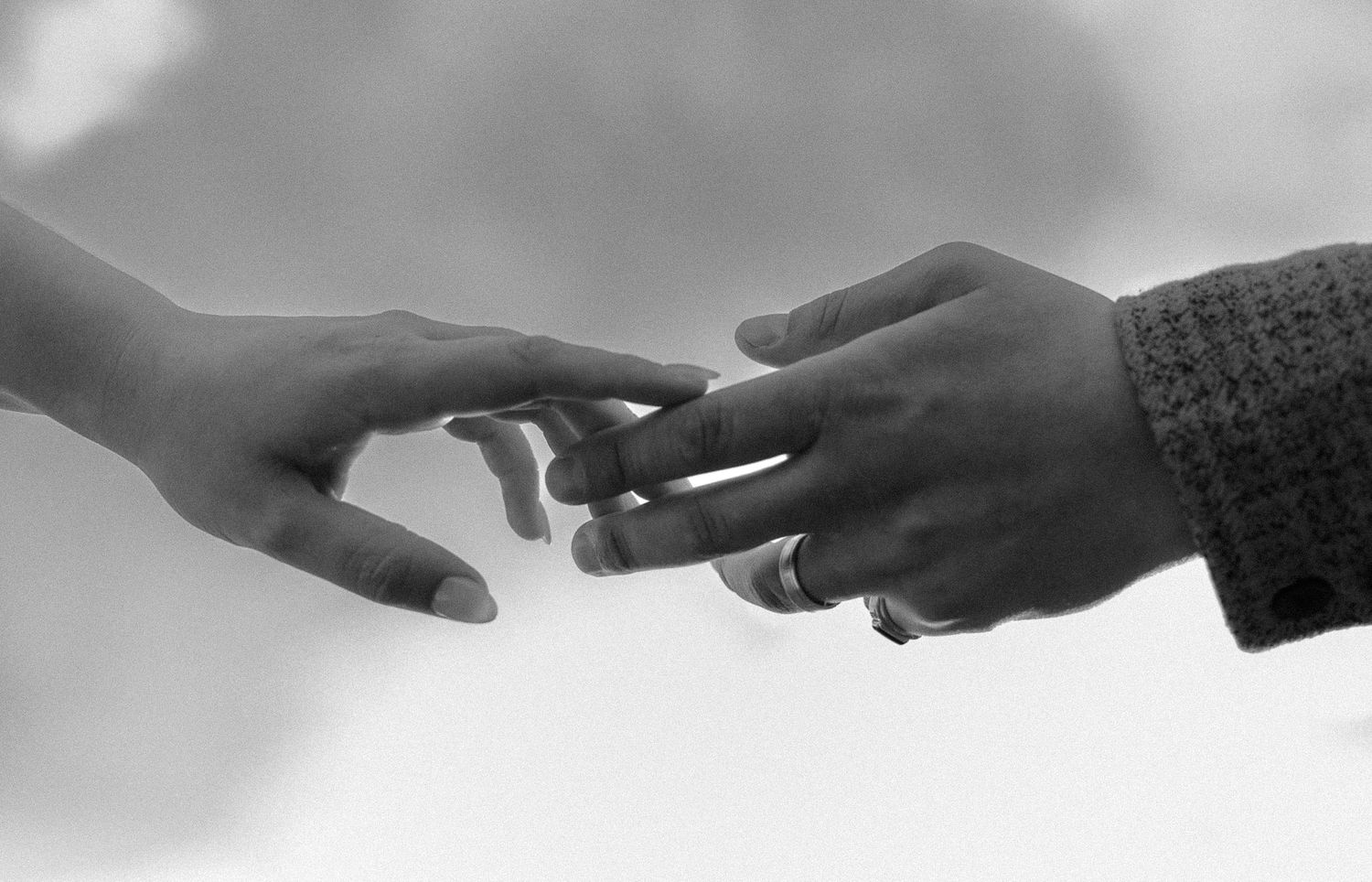 Black and white close-up of two hands reaching toward each other against a cloudy sky.