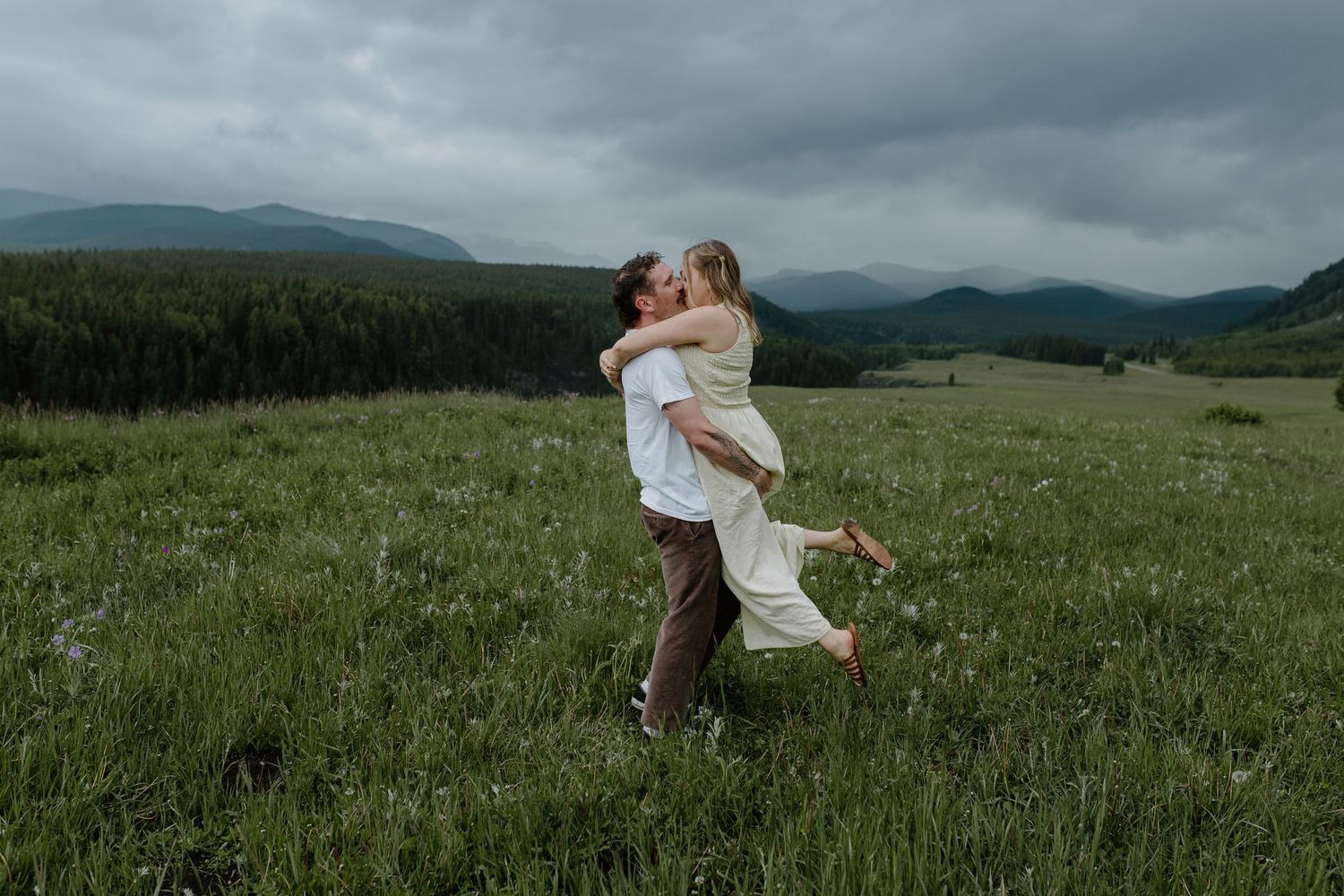 Couple embracing and kissing in a green mountain meadow under dramatic cloudy skies.