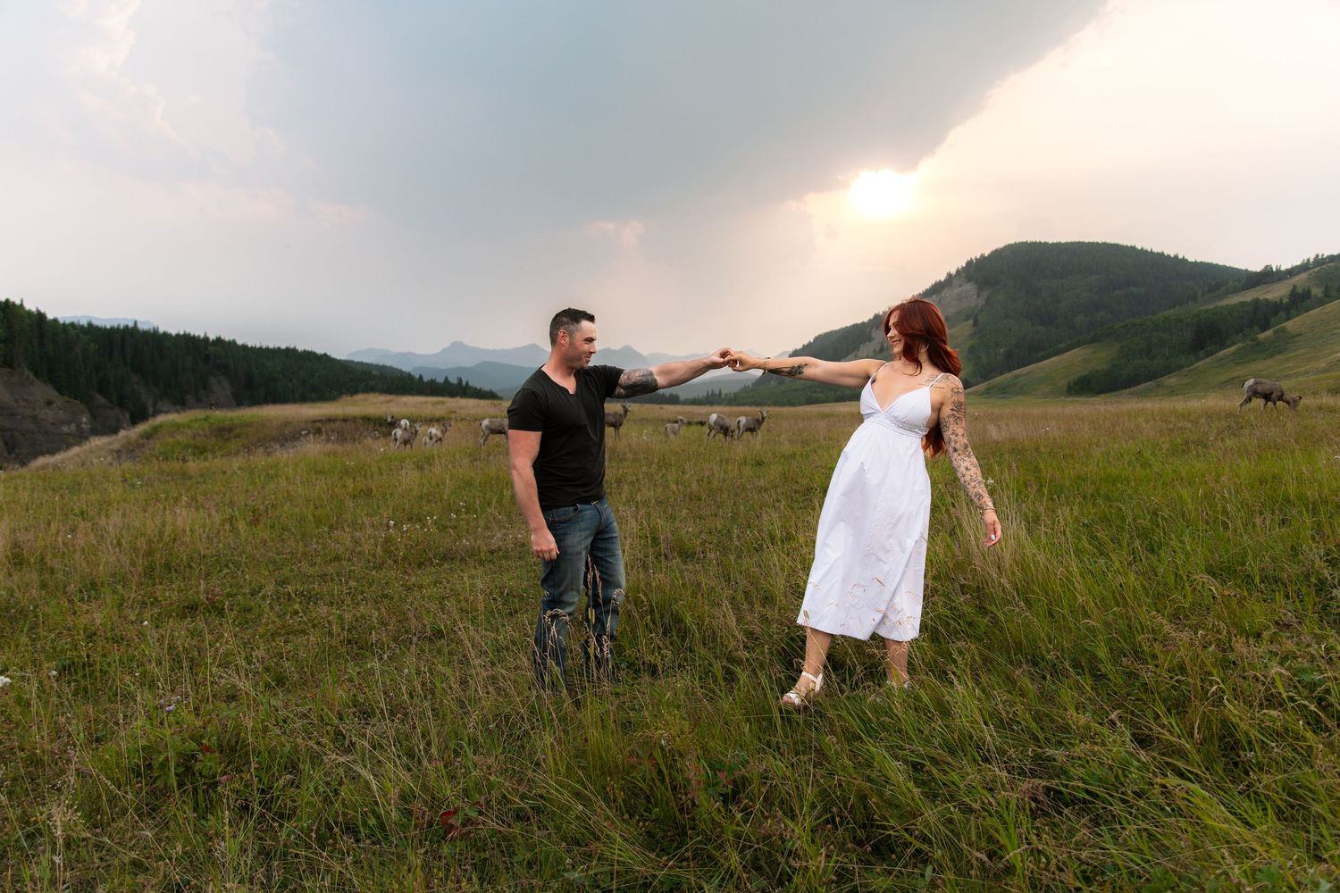 Couple holding hands in a green mountain meadow, woman in white dress, man in black shirt, cloudy sunset sky.