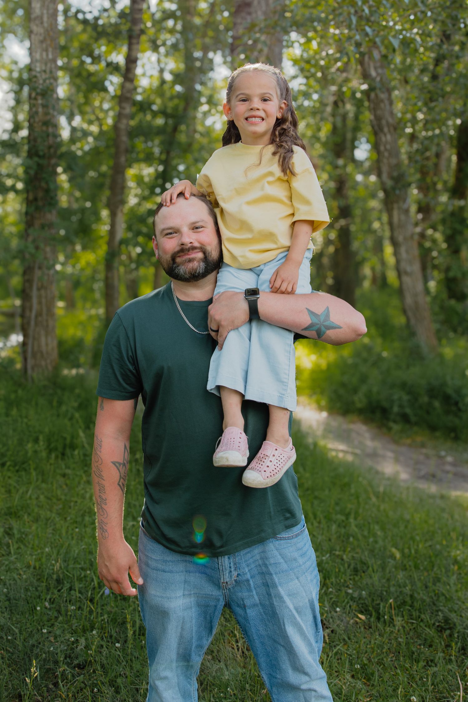 A smiling man in a green shirt carries a happy little girl on his shoulders in a lush green forest path.