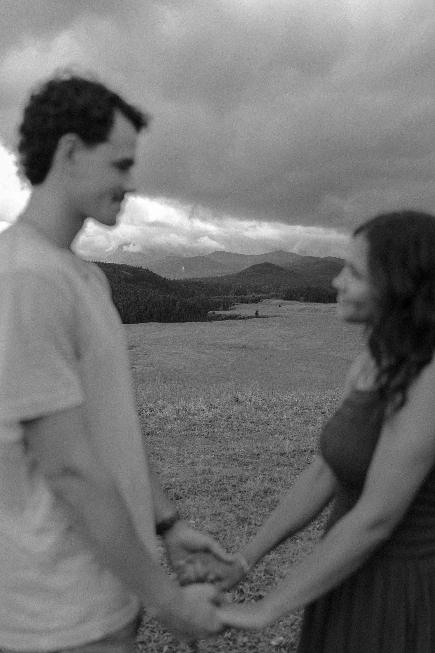 Black and white photo of a young couple holding hands in a field with mountains and dramatic clouds behind them.