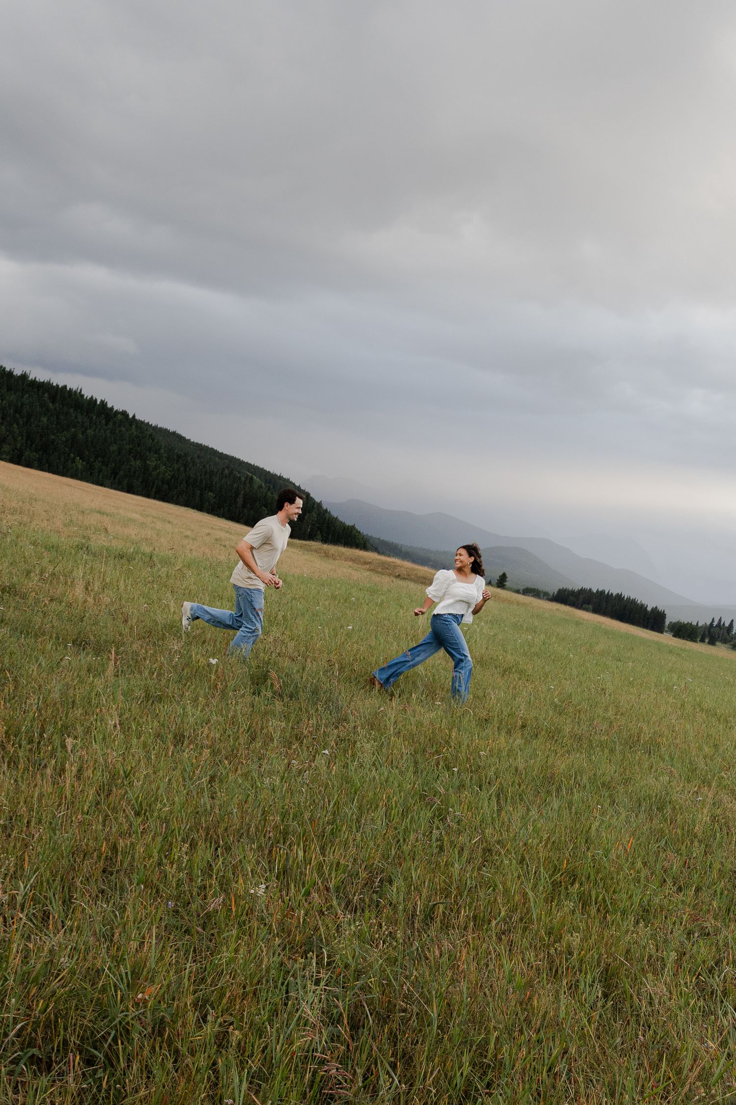 Couple running through a green meadow with mountains and forest in the background under a cloudy sky.