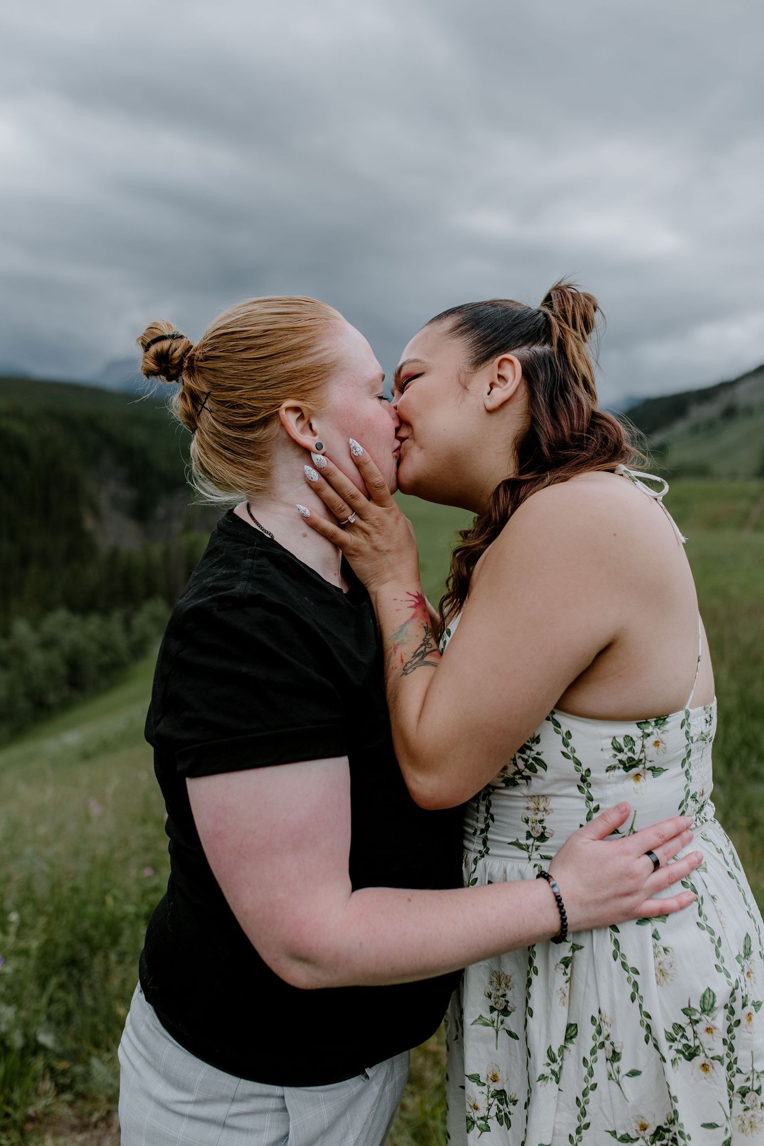 Two women sharing a romantic kiss in a lush green meadow with misty mountains in the background.