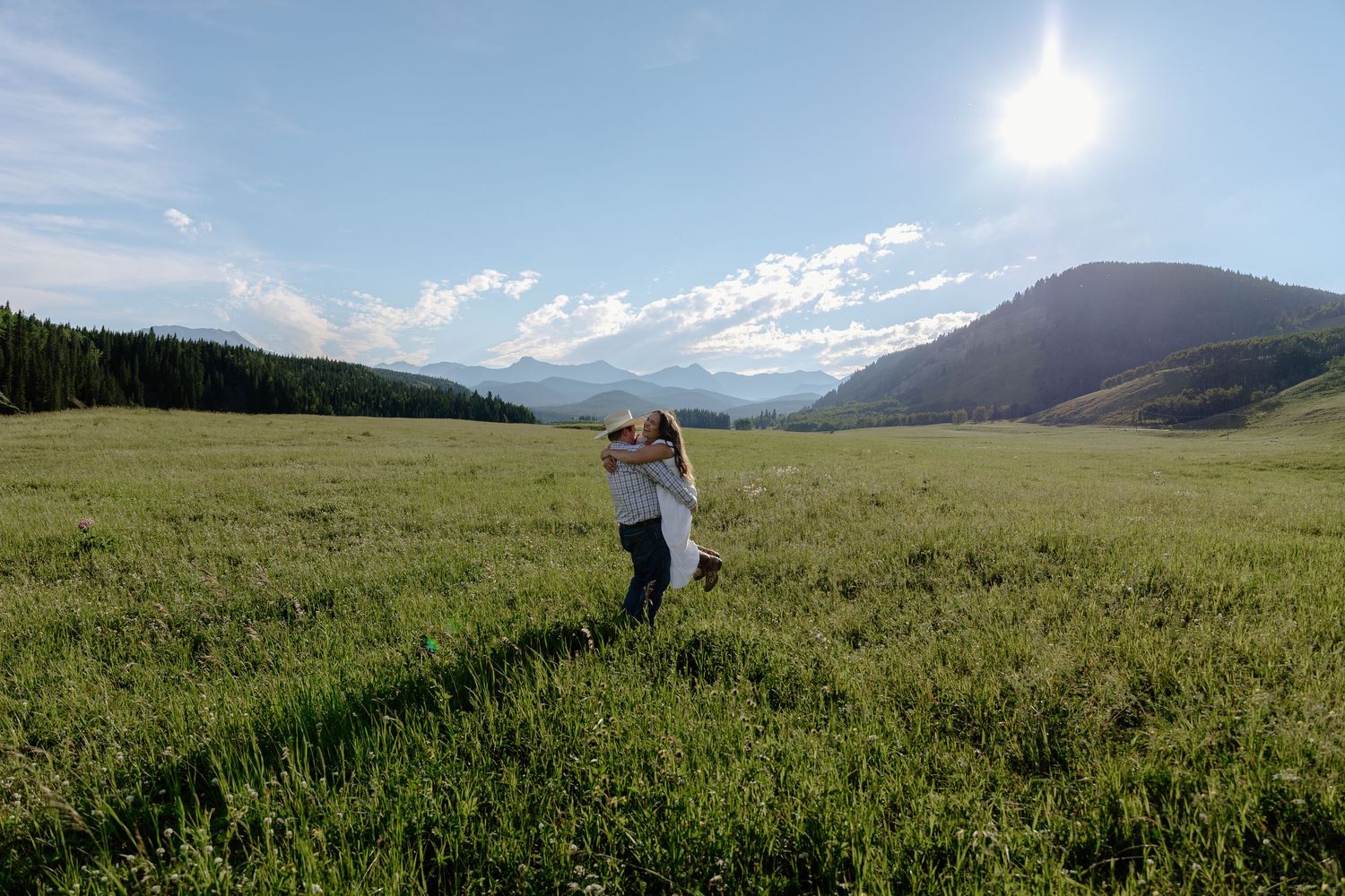 Couple embracing in a vast green meadow surrounded by mountains under a bright sunny sky.