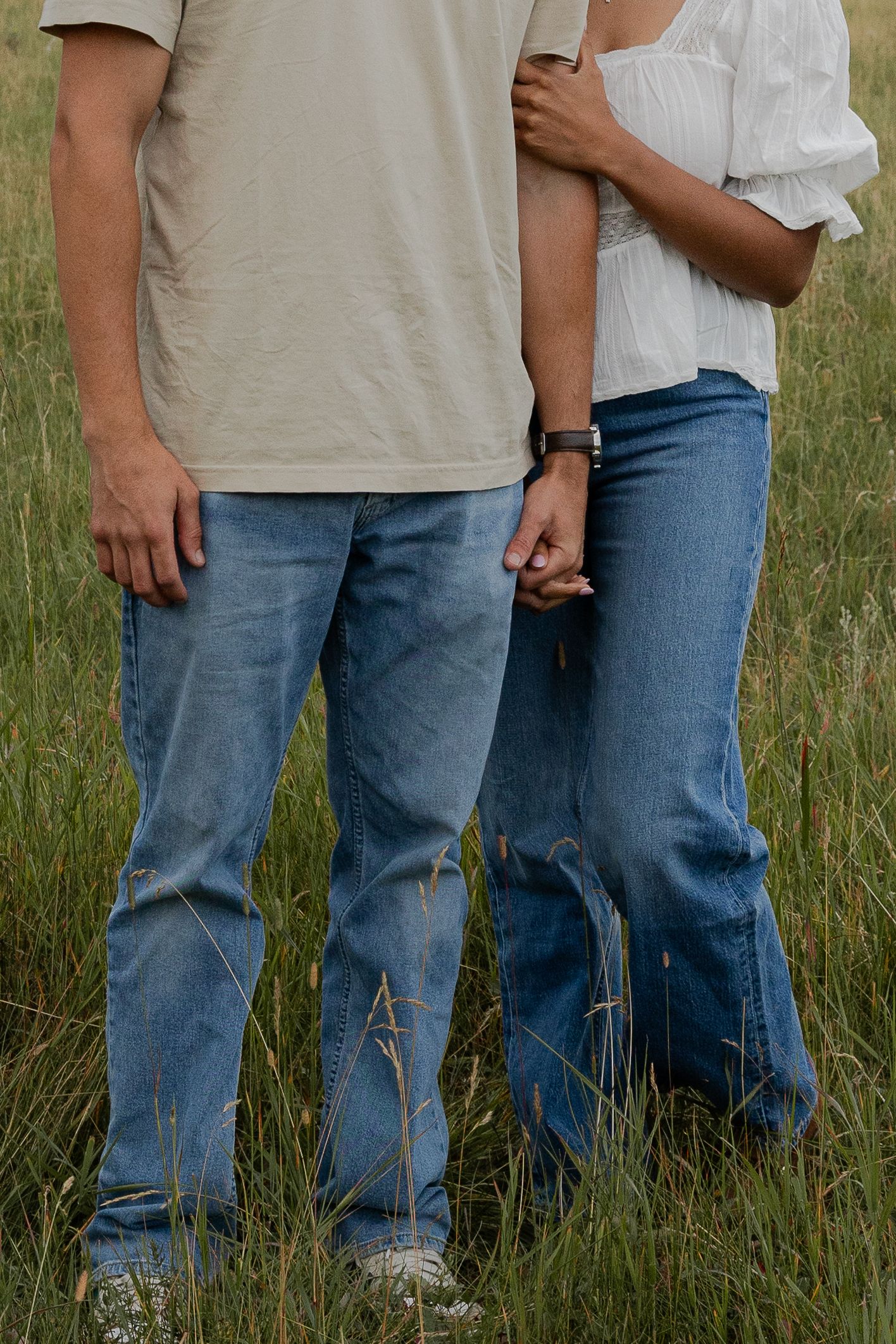 Couple standing in tall grass field, both wearing blue jeans, holding hands, casual outdoor portrait.
