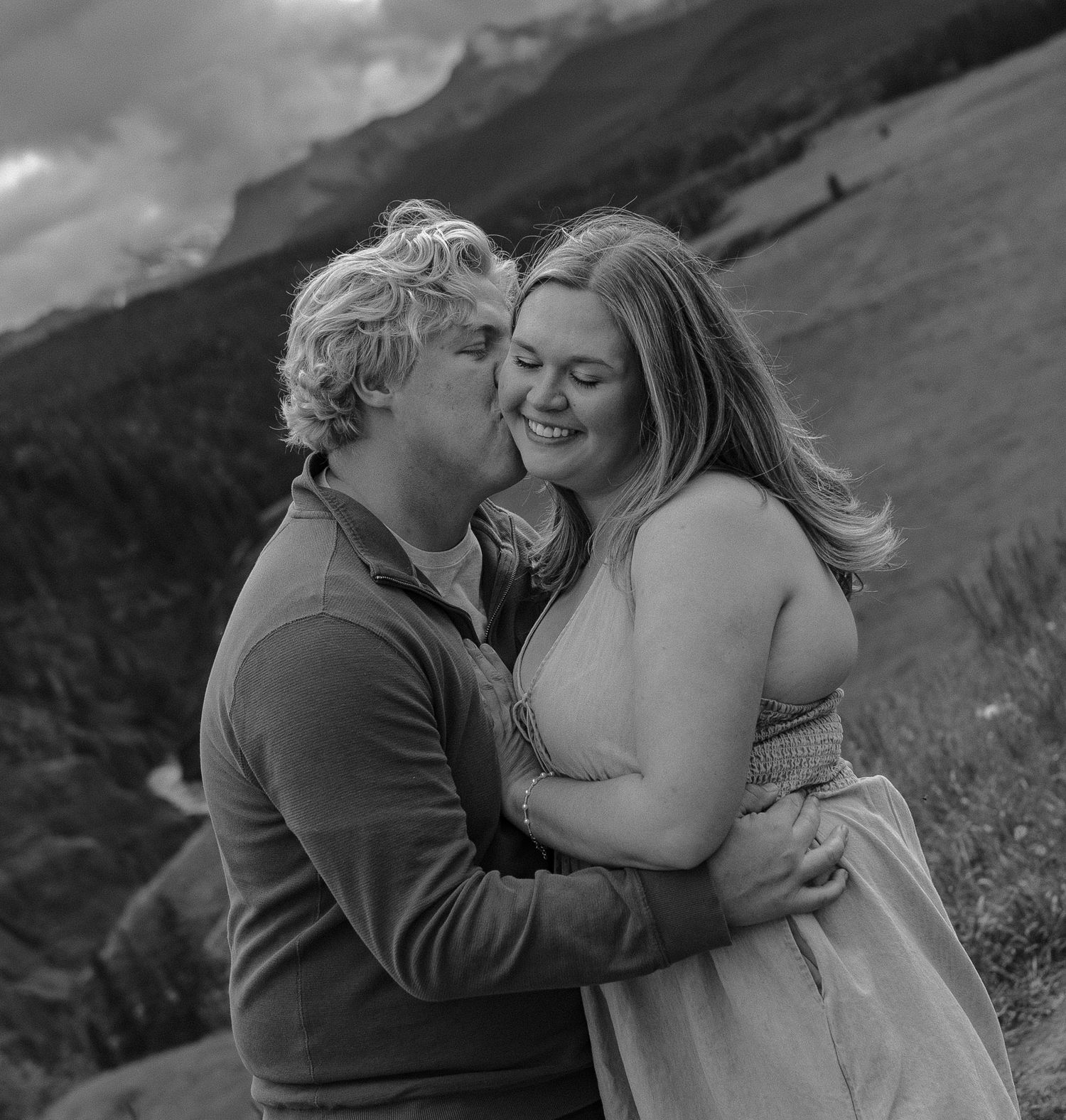 Black and white photo of a couple embracing and smiling with mountain scenery in the background.