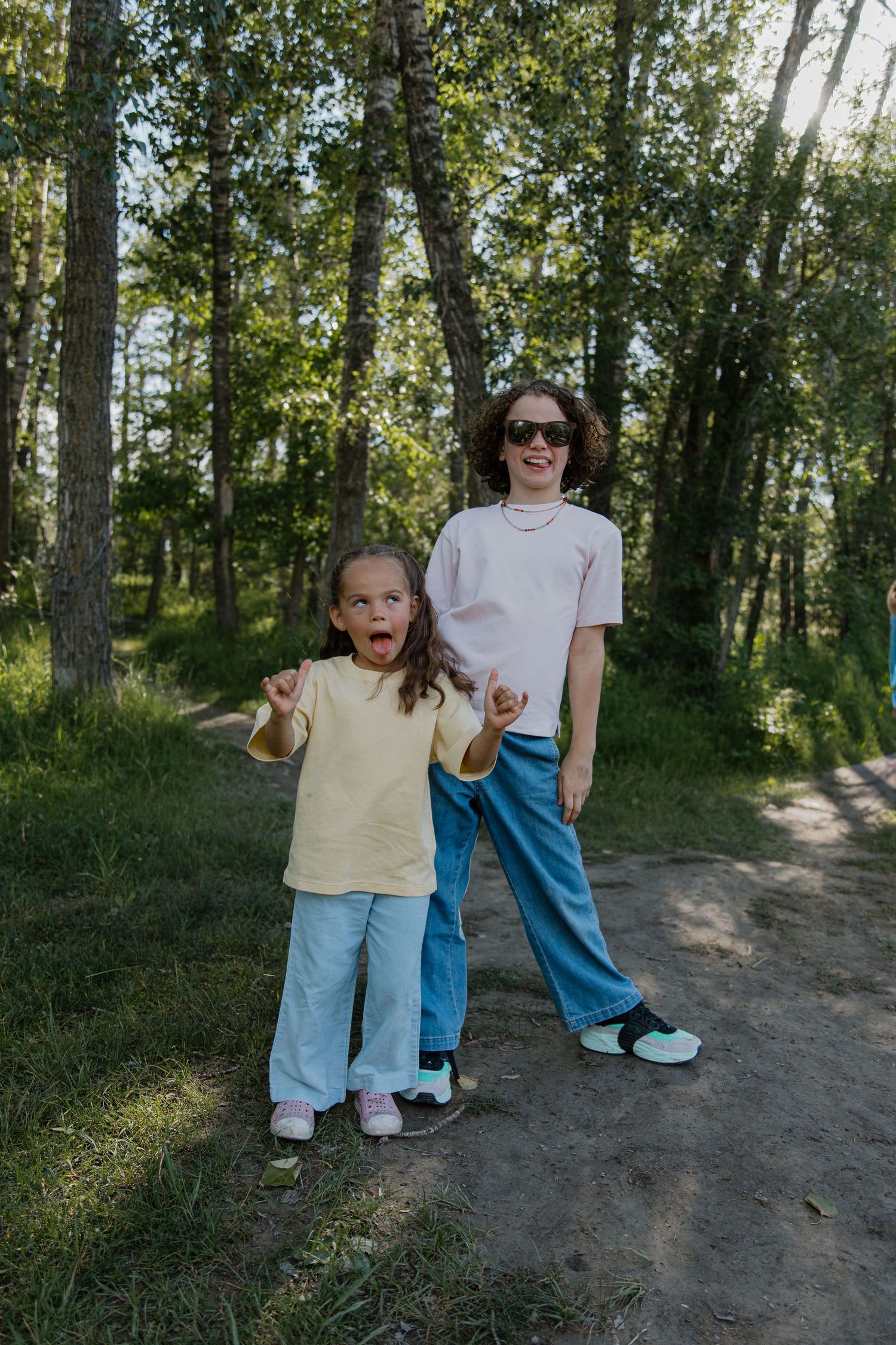 A woman and young girl in matching outfits pose on a forest path surrounded by green trees on a sunny day.