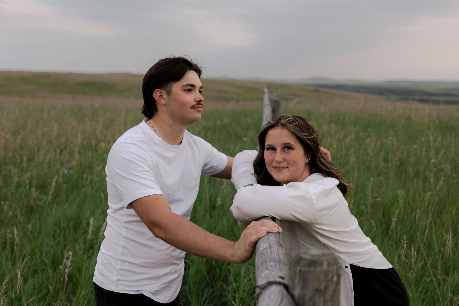 A young man and woman in white t-shirts lean on a wooden fence post in a lush green prairie field.