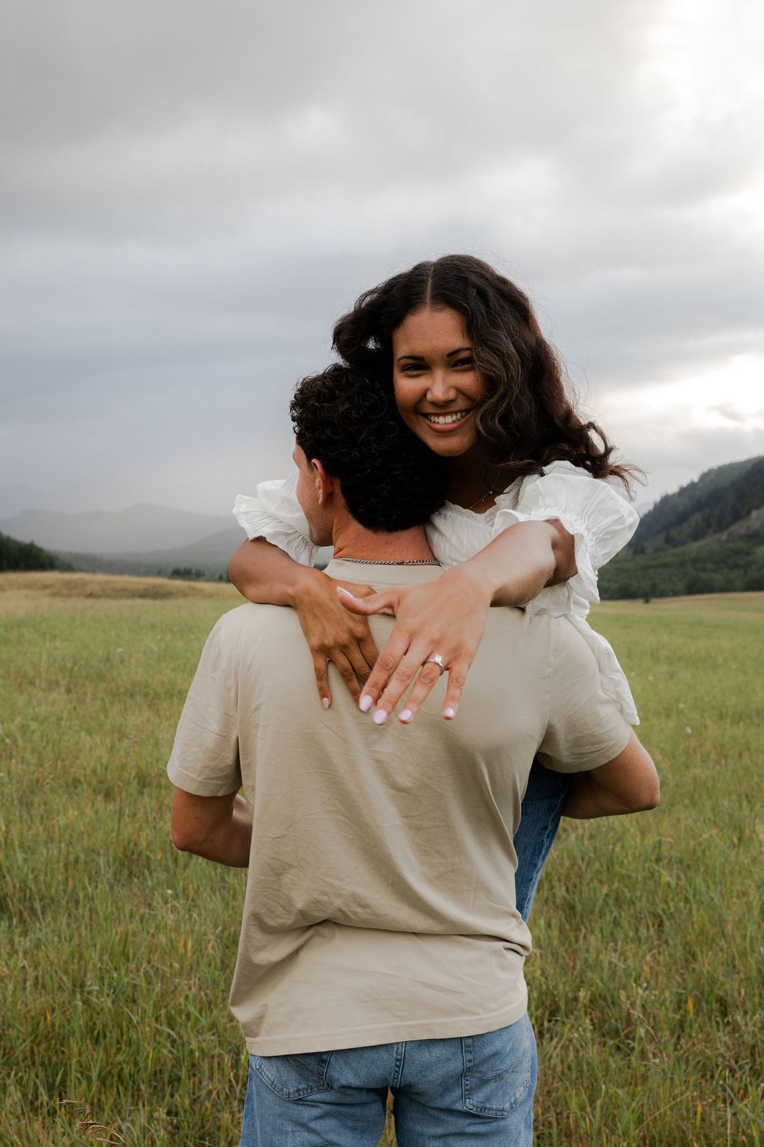 Smiling woman with engagement ring hugs man in green meadow with mountain backdrop.