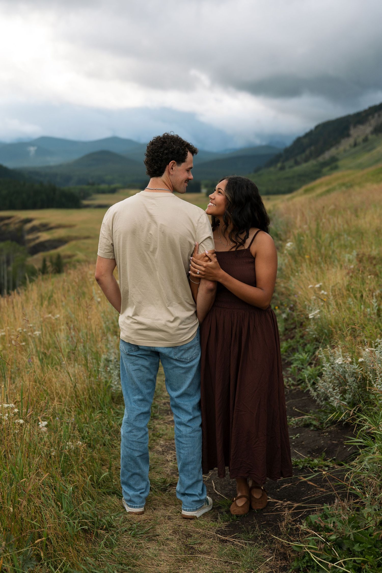 Couple embracing on a mountain meadow path, woman in brown dress smiling at man, with scenic cloudy hills behind.