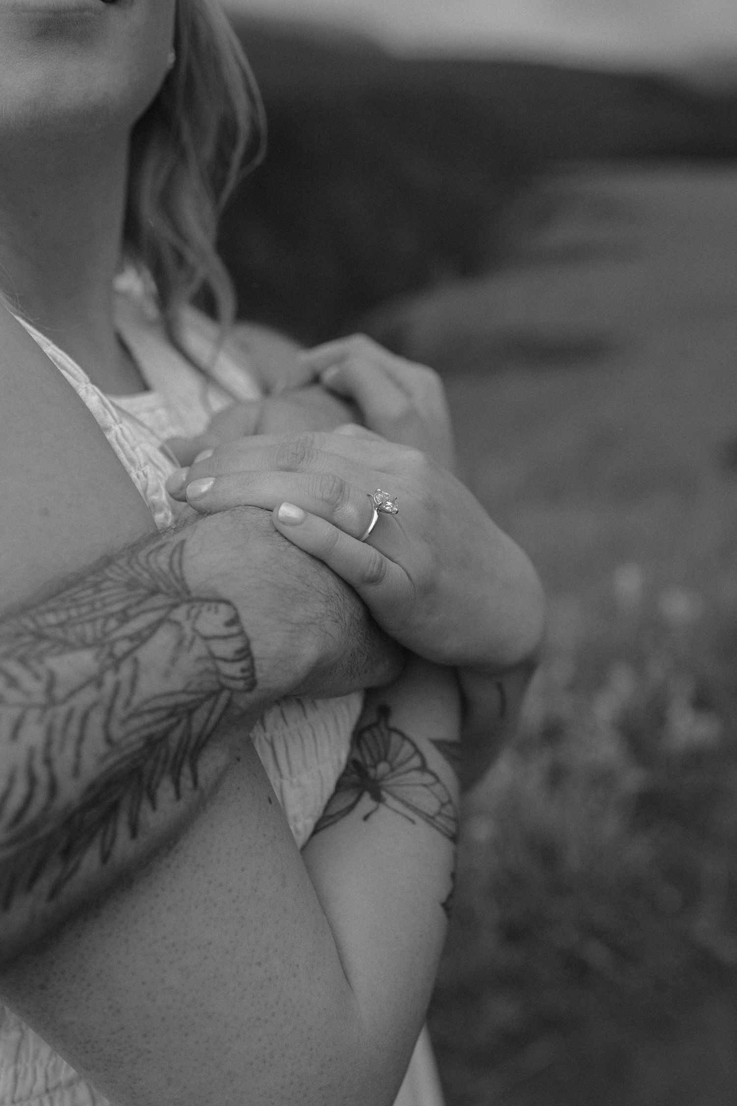 Black and white close-up of tattooed hands holding with an engagement ring visible.