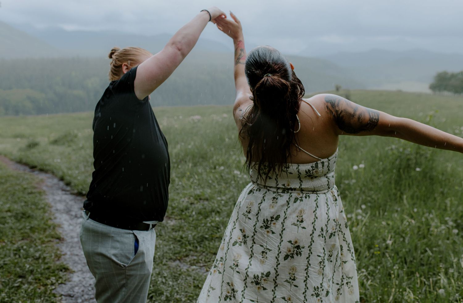 Two women dancing together on a misty green meadow path with mountains in the background.