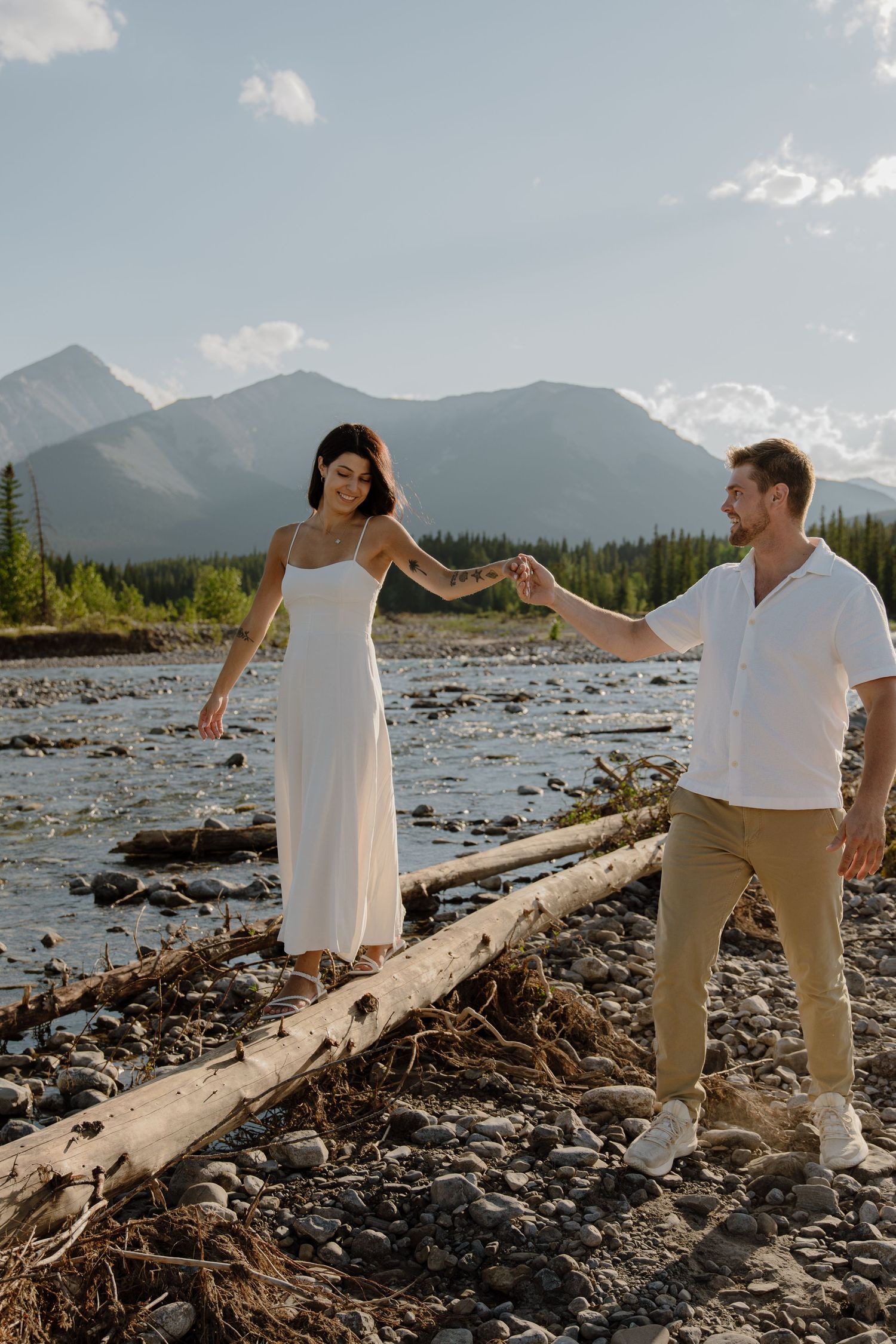 Couple holding hands on a log by a rocky river with mountain backdrop in Banff, Canada.