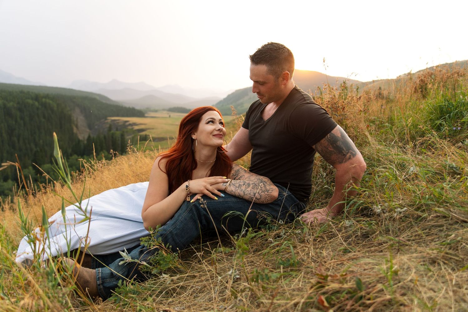 Couple sitting together on a grassy hillside at sunset, with mountains and golden fields in the background.
