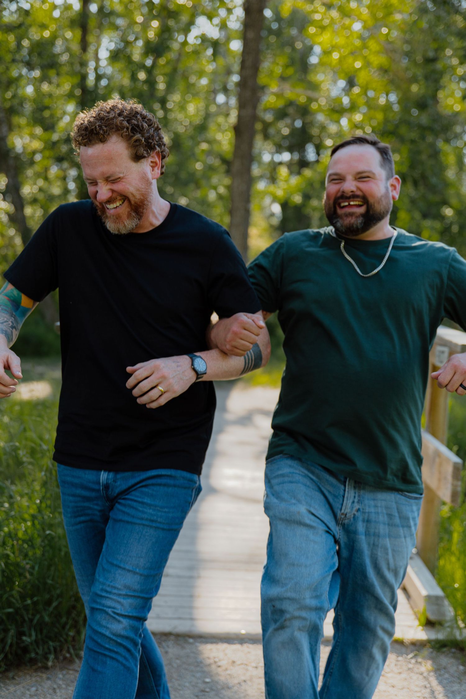 Two laughing men in casual t-shirts and jeans walk arm-in-arm along a sunny outdoor path lined with trees.