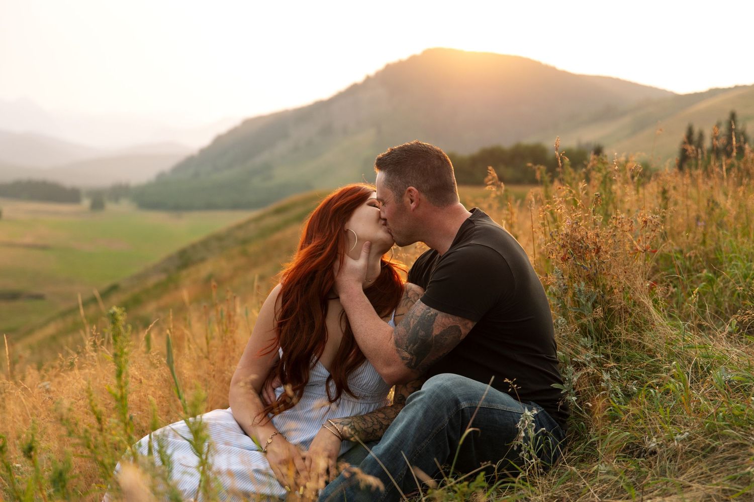 Couple sharing a romantic kiss on a grassy hillside at golden sunset with mountain landscape in background.