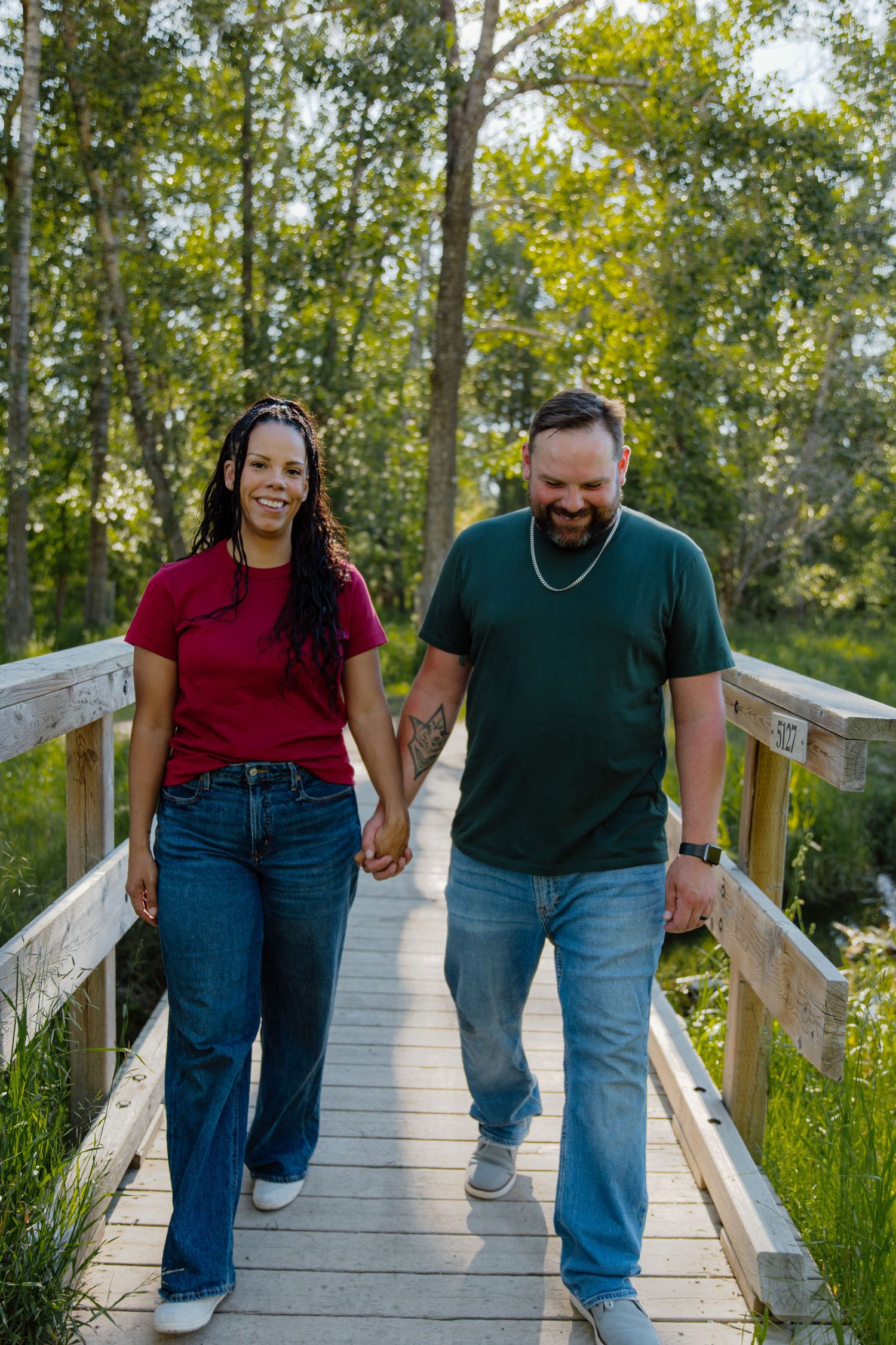 A smiling interracial couple holds hands while walking across a wooden bridge surrounded by lush green trees.