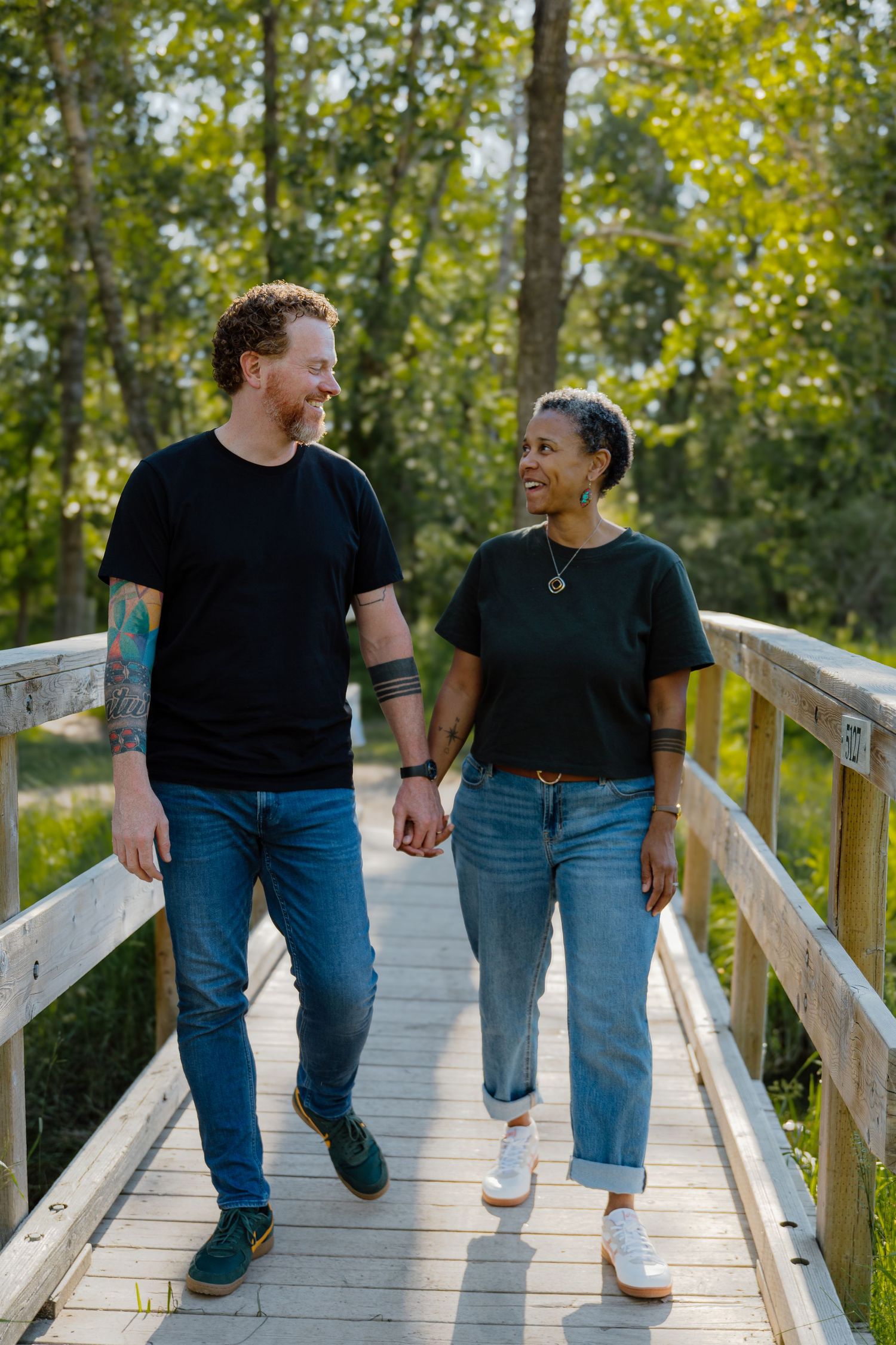 A smiling interracial couple holding hands and walking together on a wooden bridge surrounded by lush green trees.