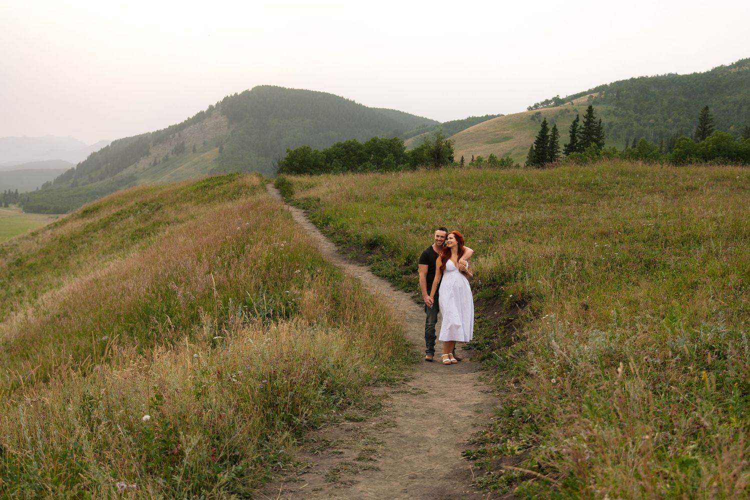 Two women stand on a scenic mountain trail surrounded by lush green meadows and rolling hills at dusk.