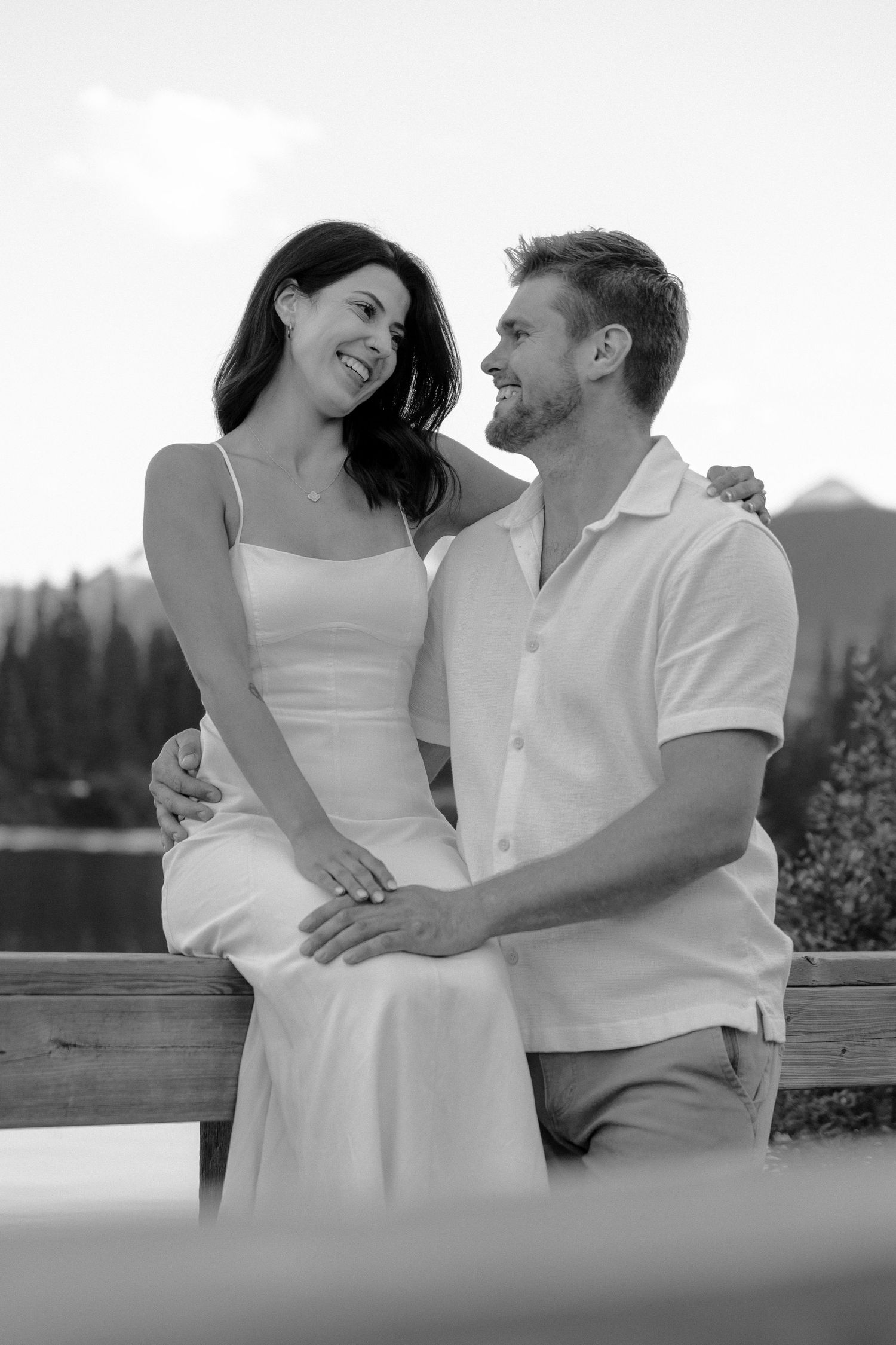 Happy couple laughing together on a wooden bench with mountain lake backdrop in black and white photo.