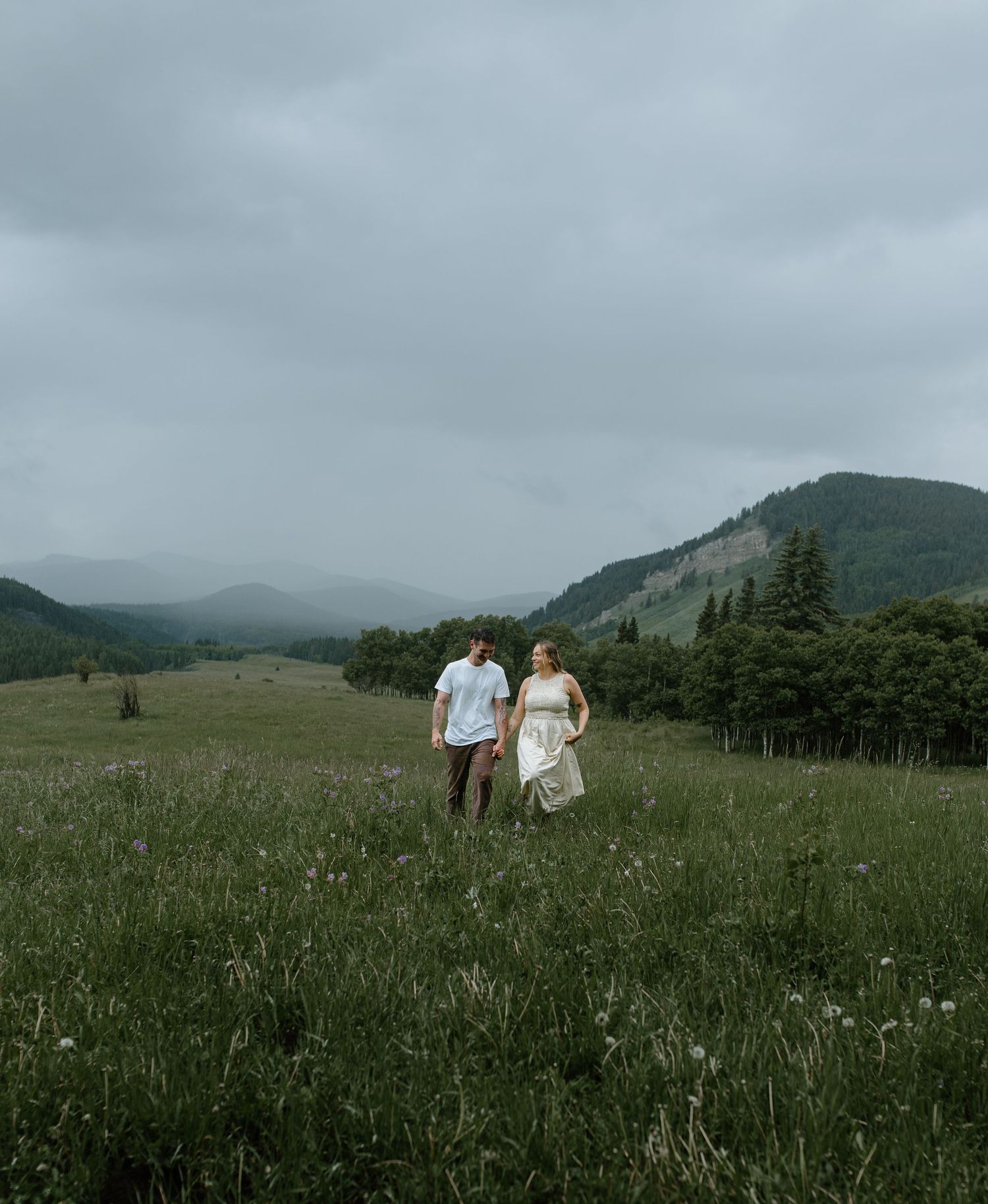 Couple walking hand in hand through a lush green mountain meadow on overcast day.