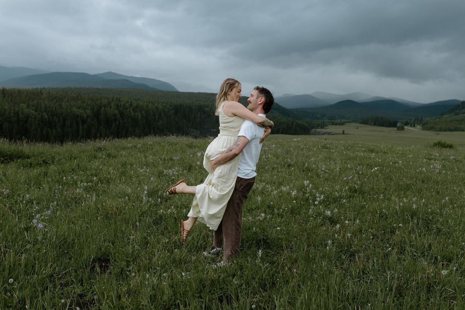 Couple embracing in a green mountain meadow under cloudy skies, man lifting woman in cream dress.