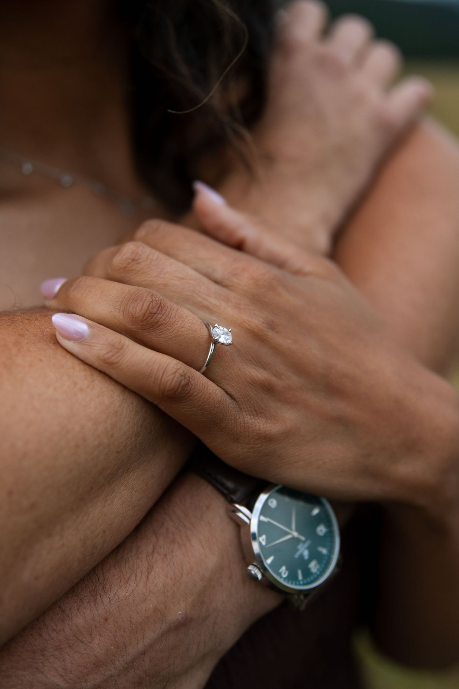 Close-up of an engagement ring with oval diamond on woman's hand resting on man's arm wearing a watch.