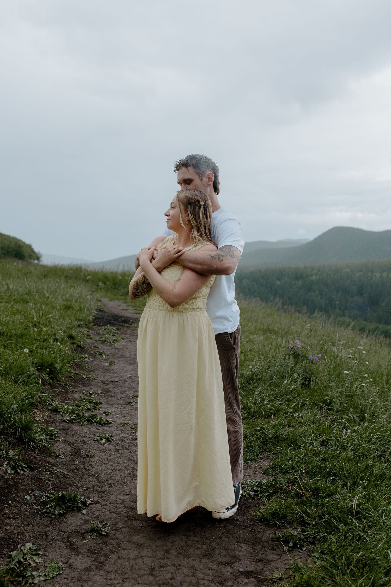 Couple embracing on a mountain trail, woman in yellow dress, man in white shirt, with green hills and cloudy sky.